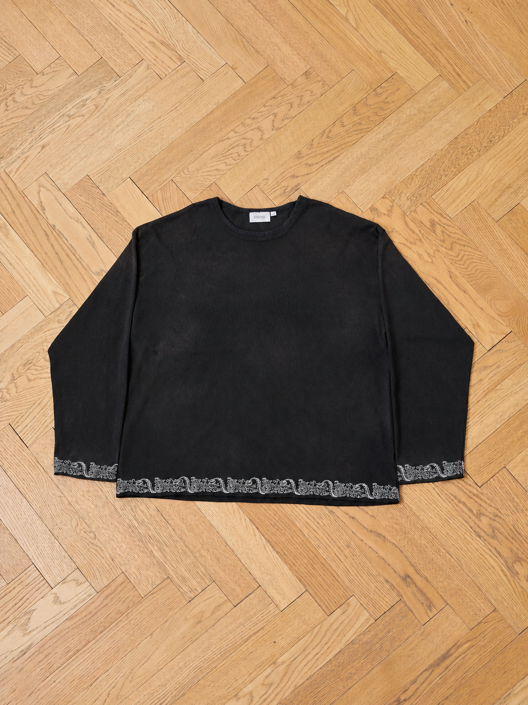 The FOUND Metallic Long Sleeve Embroidered Tee, a vintage black unisex relaxed-fit shirt with metallic embroidered trim at the hem and cuffs, is laid flat on a light wood herringbone floor.