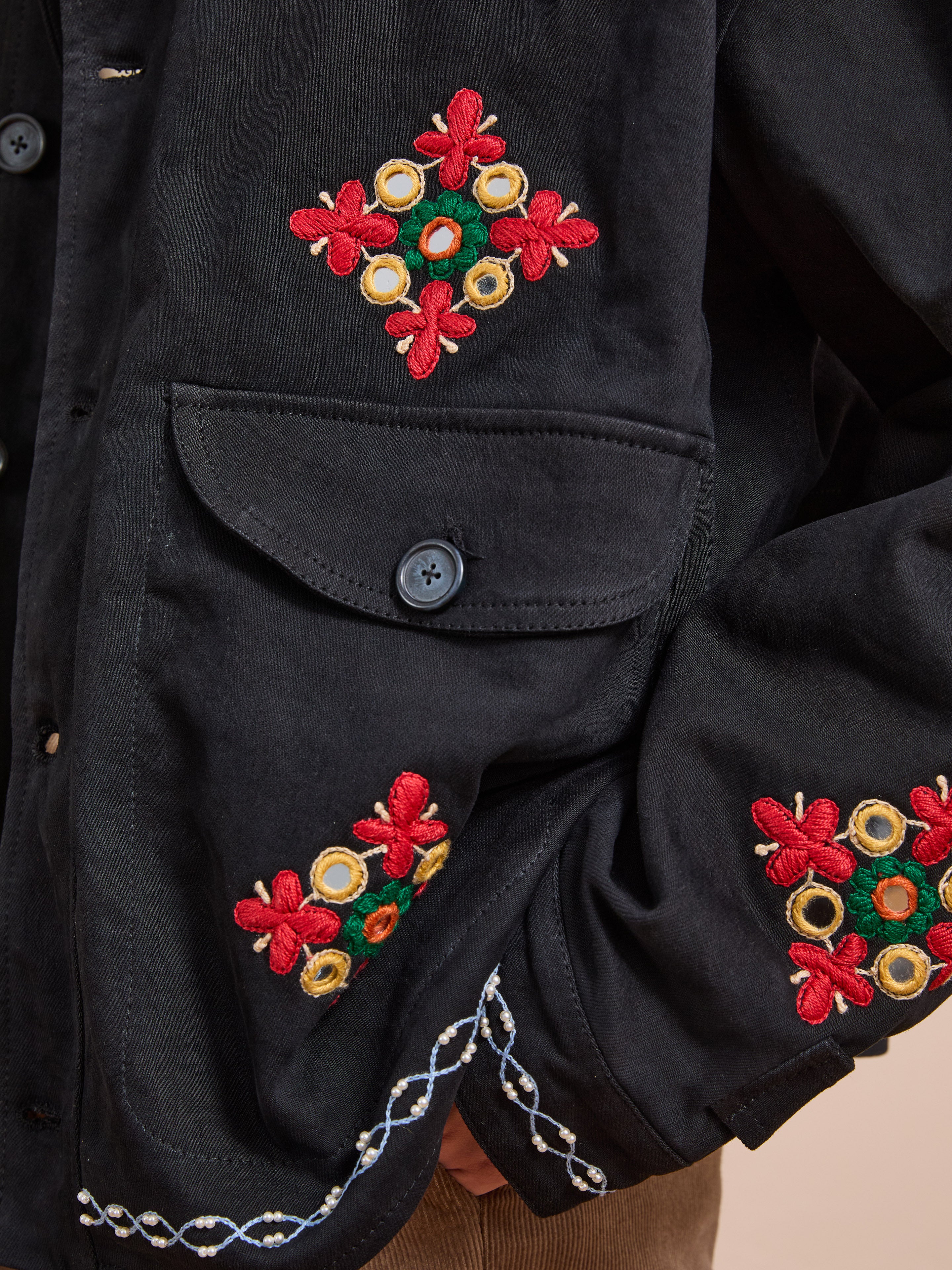Close-up of the Mirror Ornament Hand Embroidered Work Jacket by Found, handcrafted with red, yellow, and green floral Aari embroidery and finished with a white decorative trim near the cuff.
