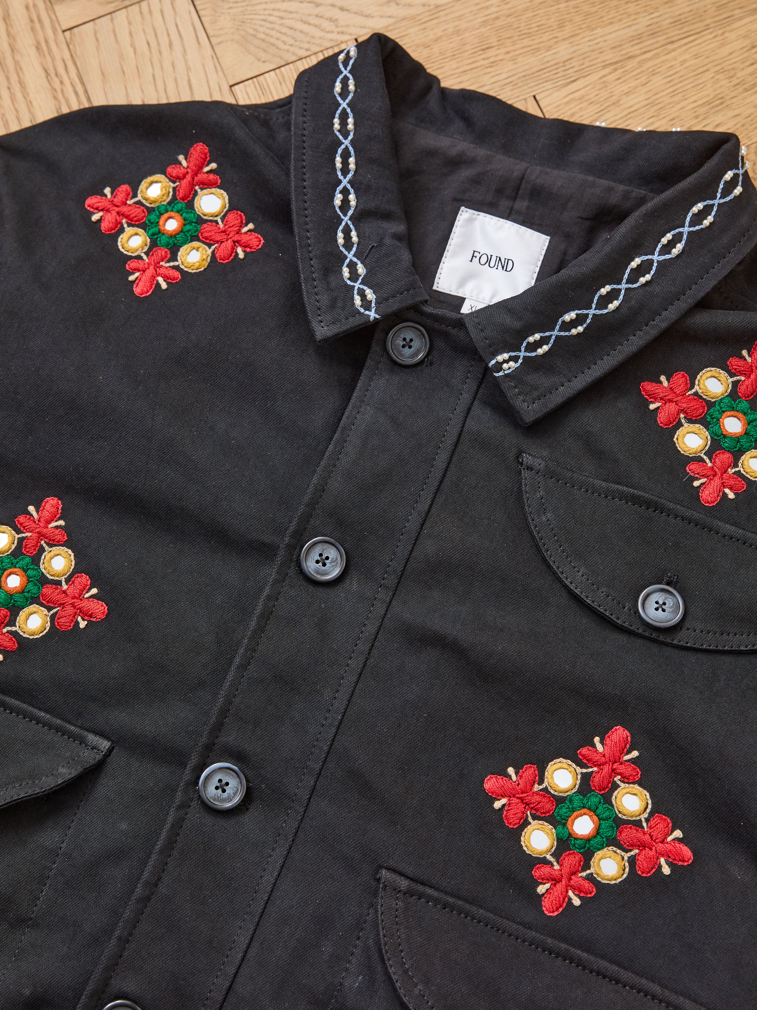 A black Mirror Ornament Hand Embroidered Work Jacket from Found, featuring Aari-inspired red, yellow, and green floral embroidery and white collar stitching, is displayed on a wooden surface.