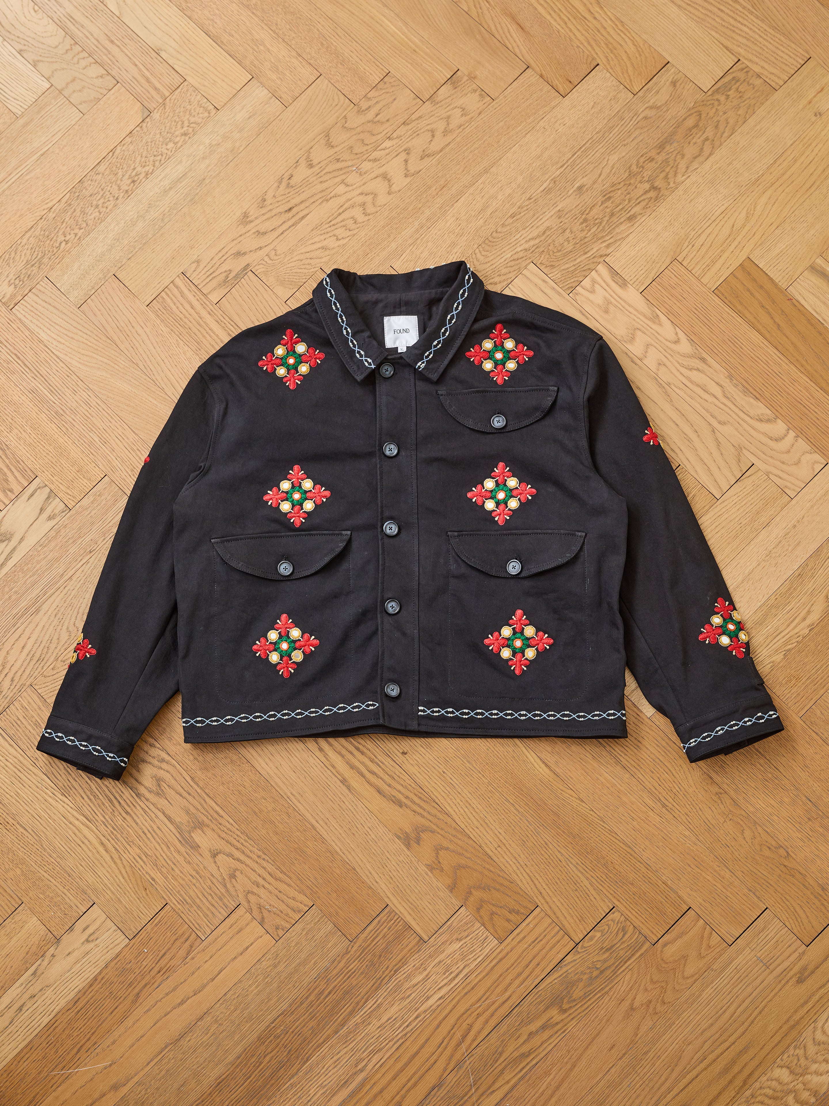 The Found Mirror Ornament Hand Embroidered Work Jacket is a black handcrafted cotton piece featuring two front pockets, vivid red and green floral Aari embroidery, and chain-like trim, shown flat on a wooden herringbone floor.
