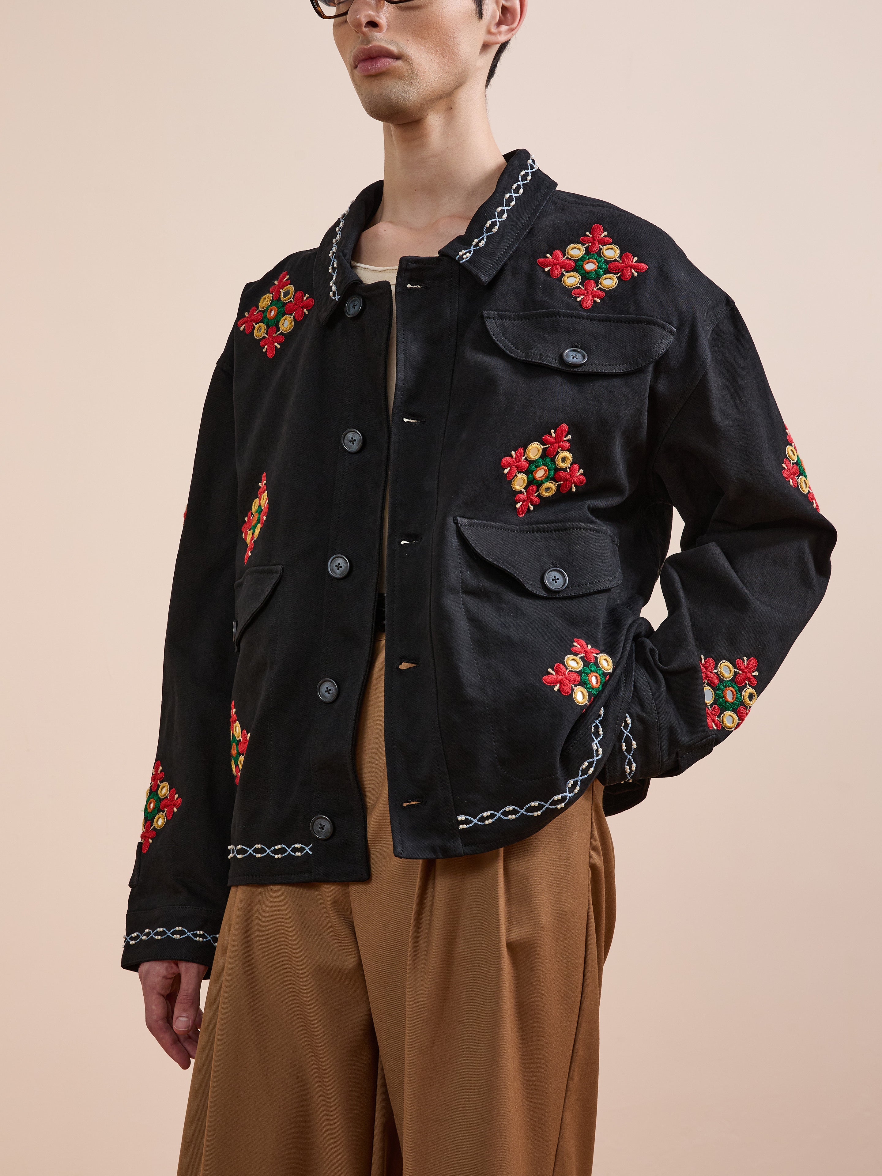 A person wearing the Found Mirror Ornament Hand Embroidered Work Jacket—black with red and green floral details—and tan pants, stands against a plain beige background.