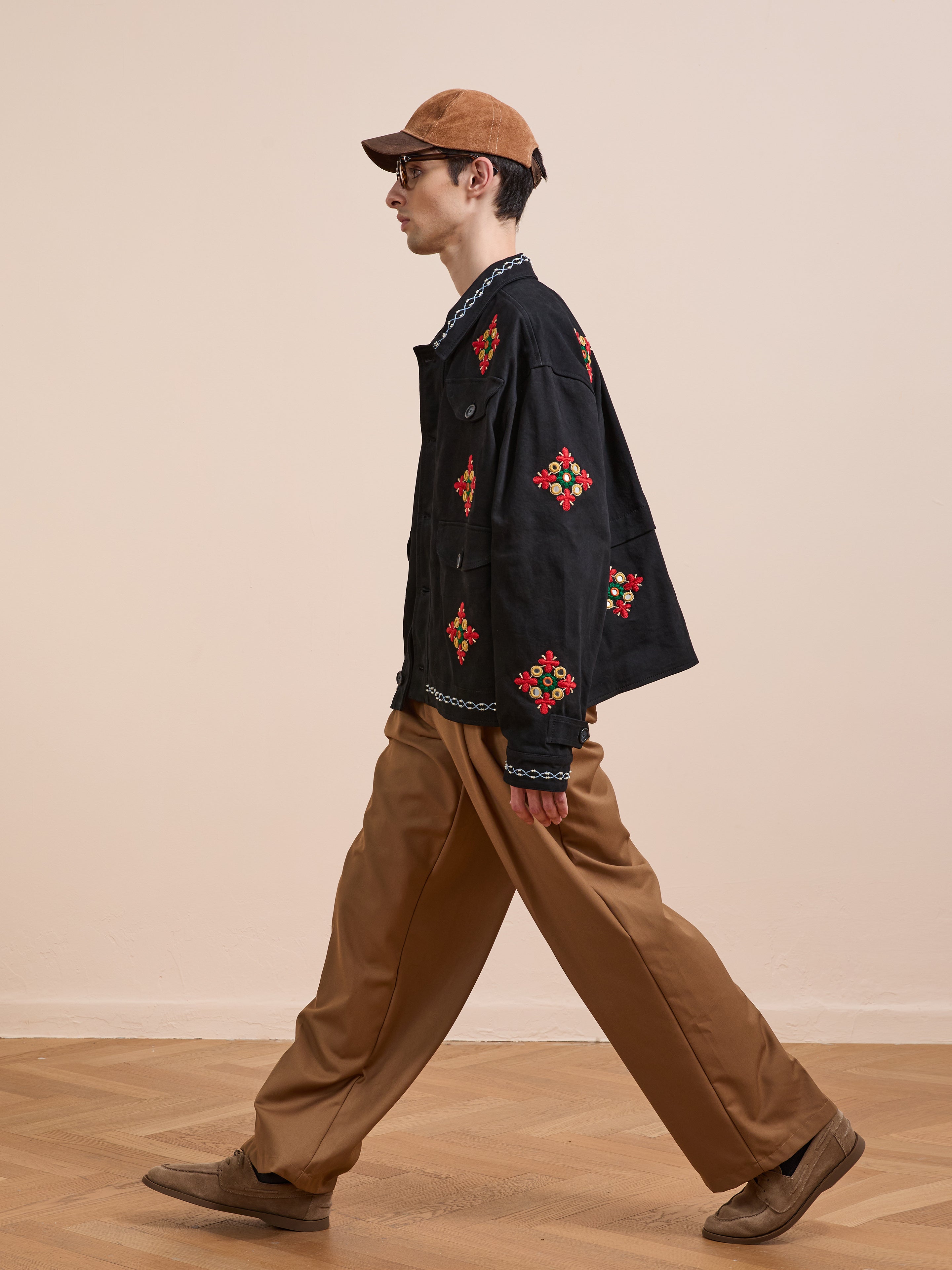 A man in the Found Mirror Ornament Hand Embroidered Work Jacket, a brown cap, tan wide-leg pants, and brown shoes walks indoors on a wooden floor.