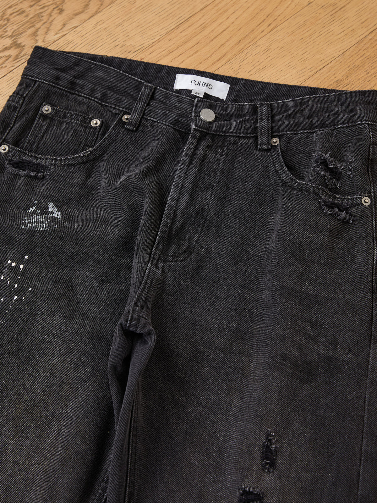 Monet Paint Jeans from FOUND, featuring vintage black distressed denim with silver rivets, are displayed flat on a light wooden surface.