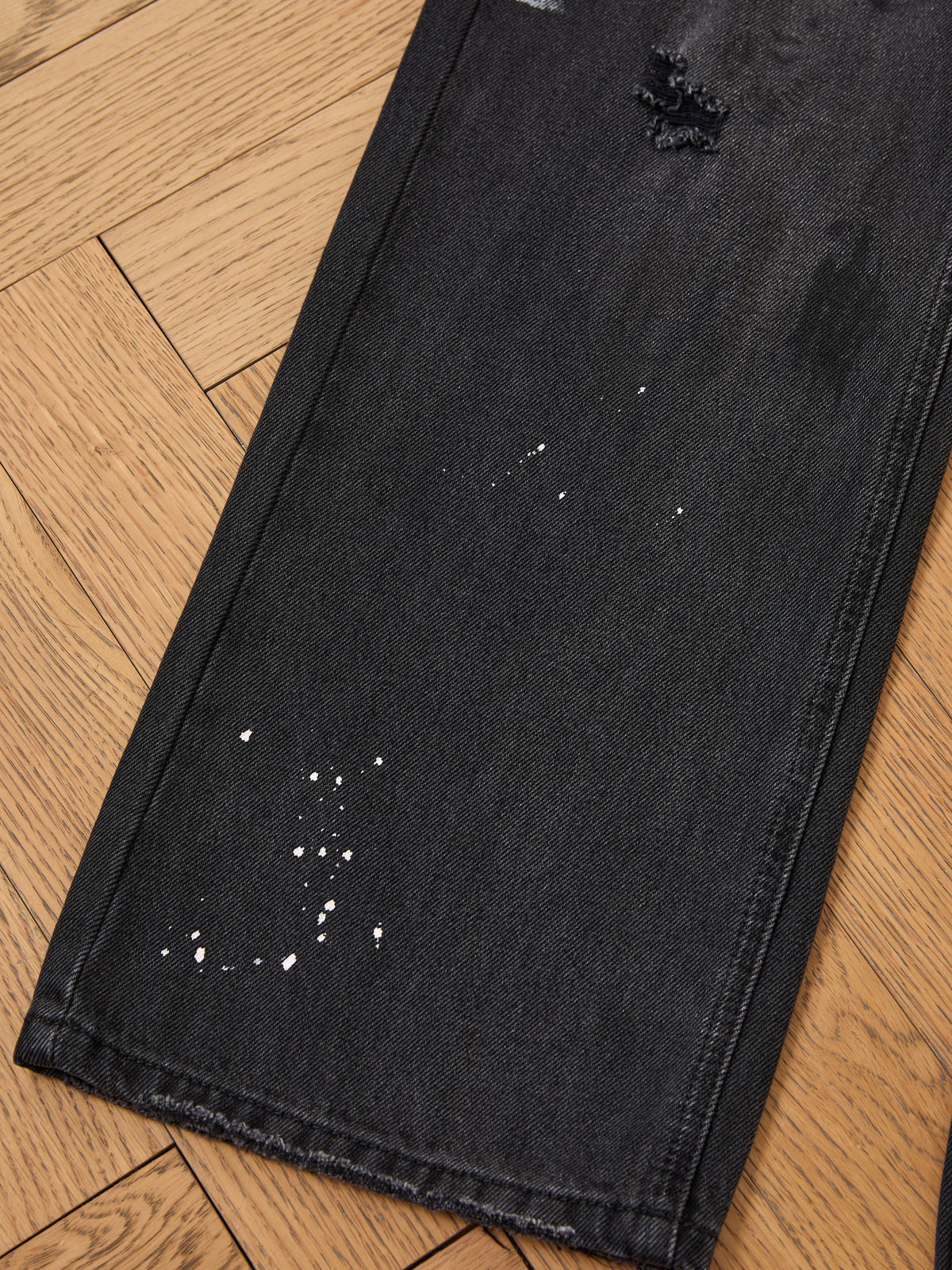 A close-up of FOUND Monet Paint Jeans with a vintage black wash and paint splatters, showing signs of wear, laid flat on a wooden floor.