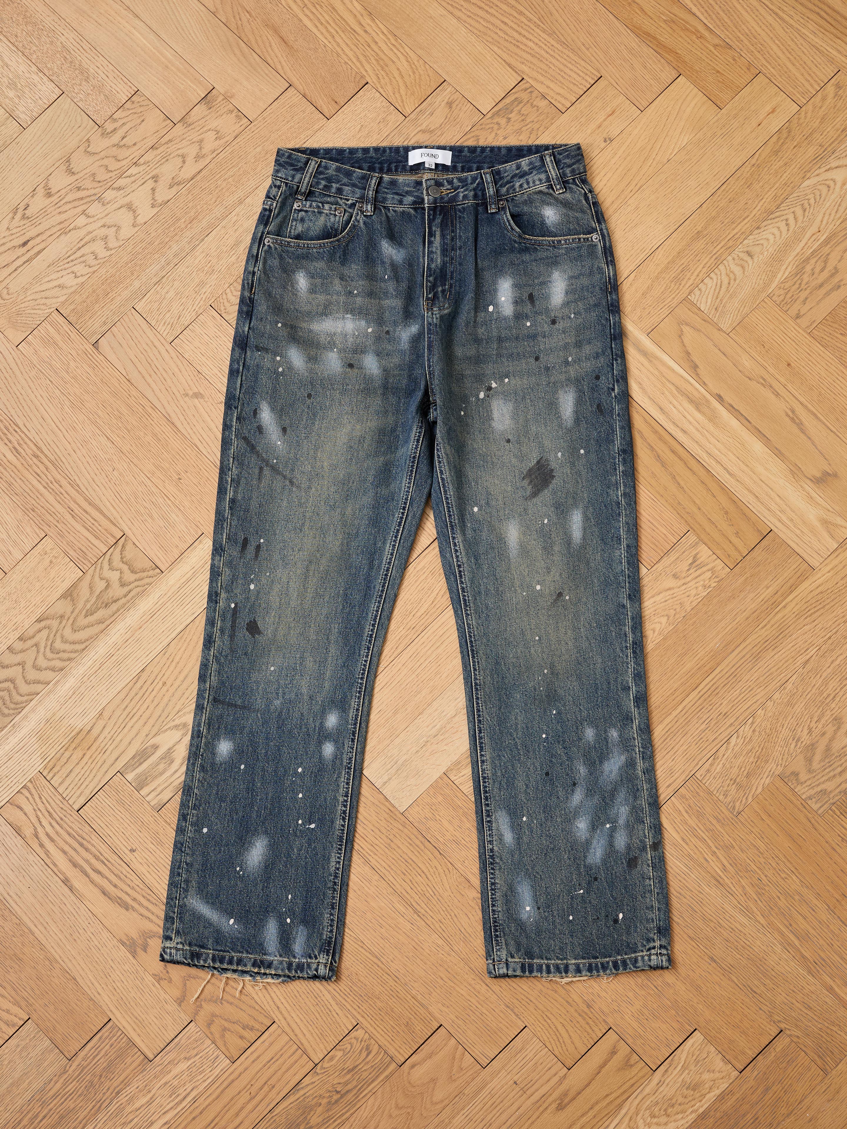 A pair of FOUND Monet Paint Jeans with a unisex relaxed fit and vintage blue wash, featuring paint splatter detail, lies flat on a light wood herringbone floor.