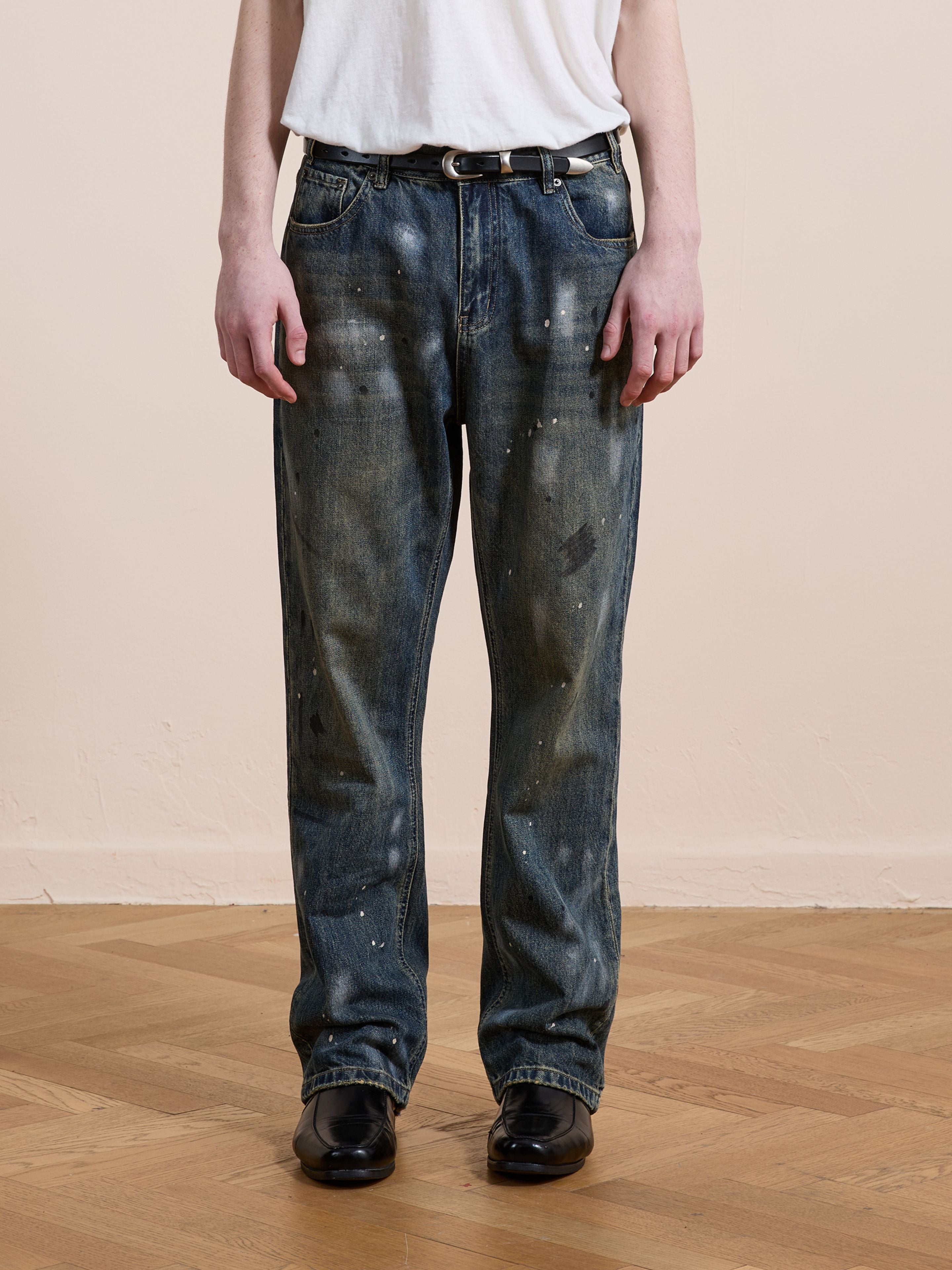 A person wearing a white t-shirt and FOUND Monet Paint Jeans in vintage blue stands on a wooden floor against a beige wall, styled with a black belt and black shoes.