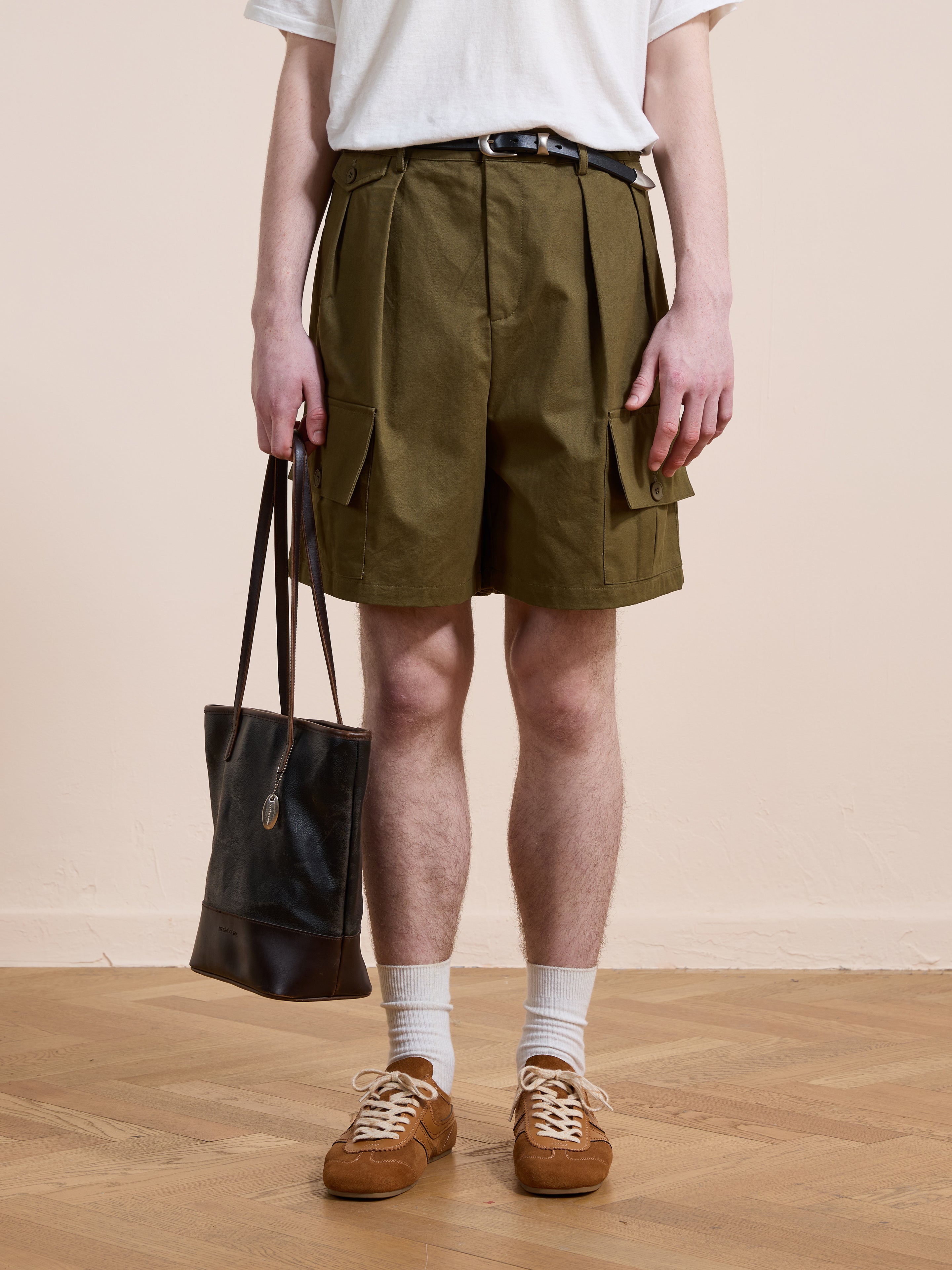 A person wears Found Multi Pocket Shorts with a white t-shirt, white socks, brown sneakers, and holds a black tote bag. The shorts’ utility design adds function to the casual outfit. They stand on a wooden floor against a beige wall.