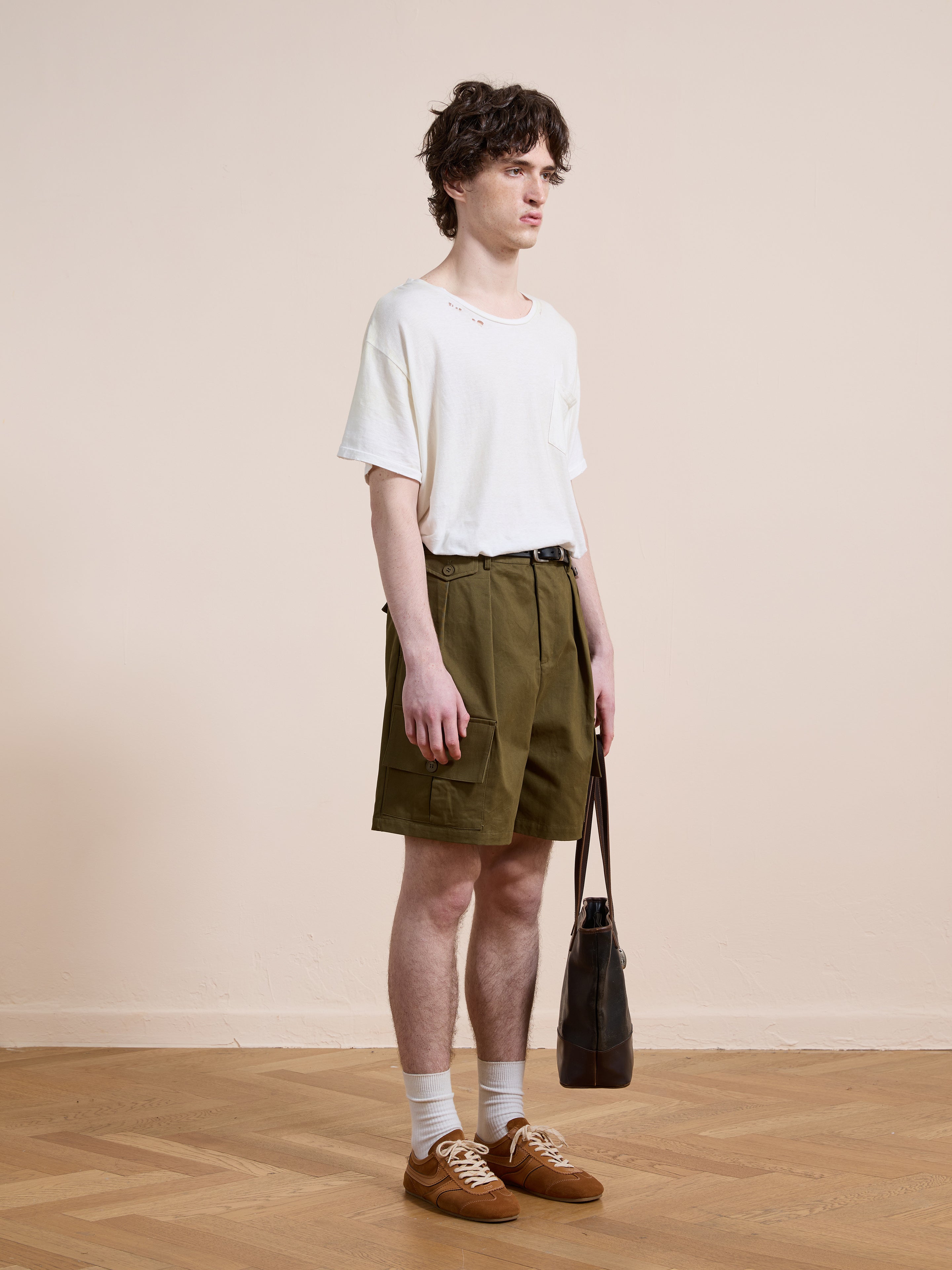 A person stands indoors against a beige wall, wearing a white t-shirt with holes, Found Multi Pocket Shorts in olive green, white socks, brown sneakers, and holding a black bag.