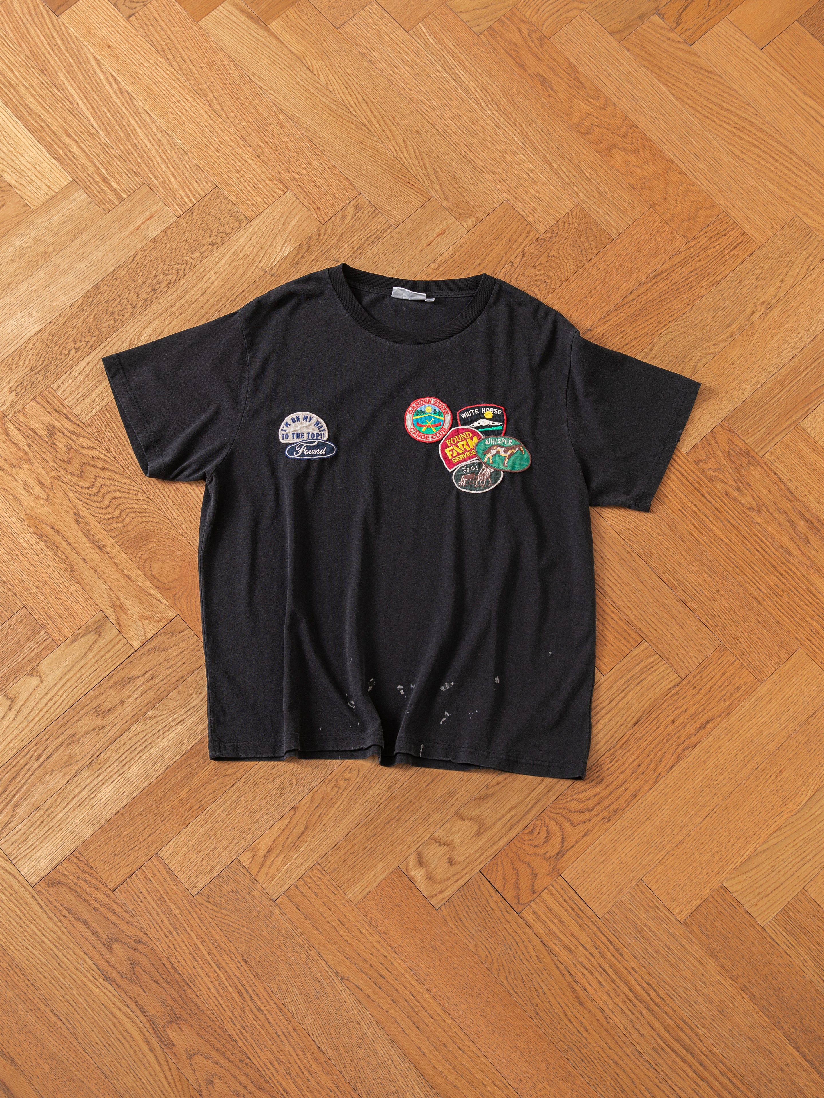 The Multi Patch Washed Tee by FOUND, featuring a black design adorned with multiple colorful patches, is displayed on a wooden parquet floor. The shirt has small holes near the bottom.