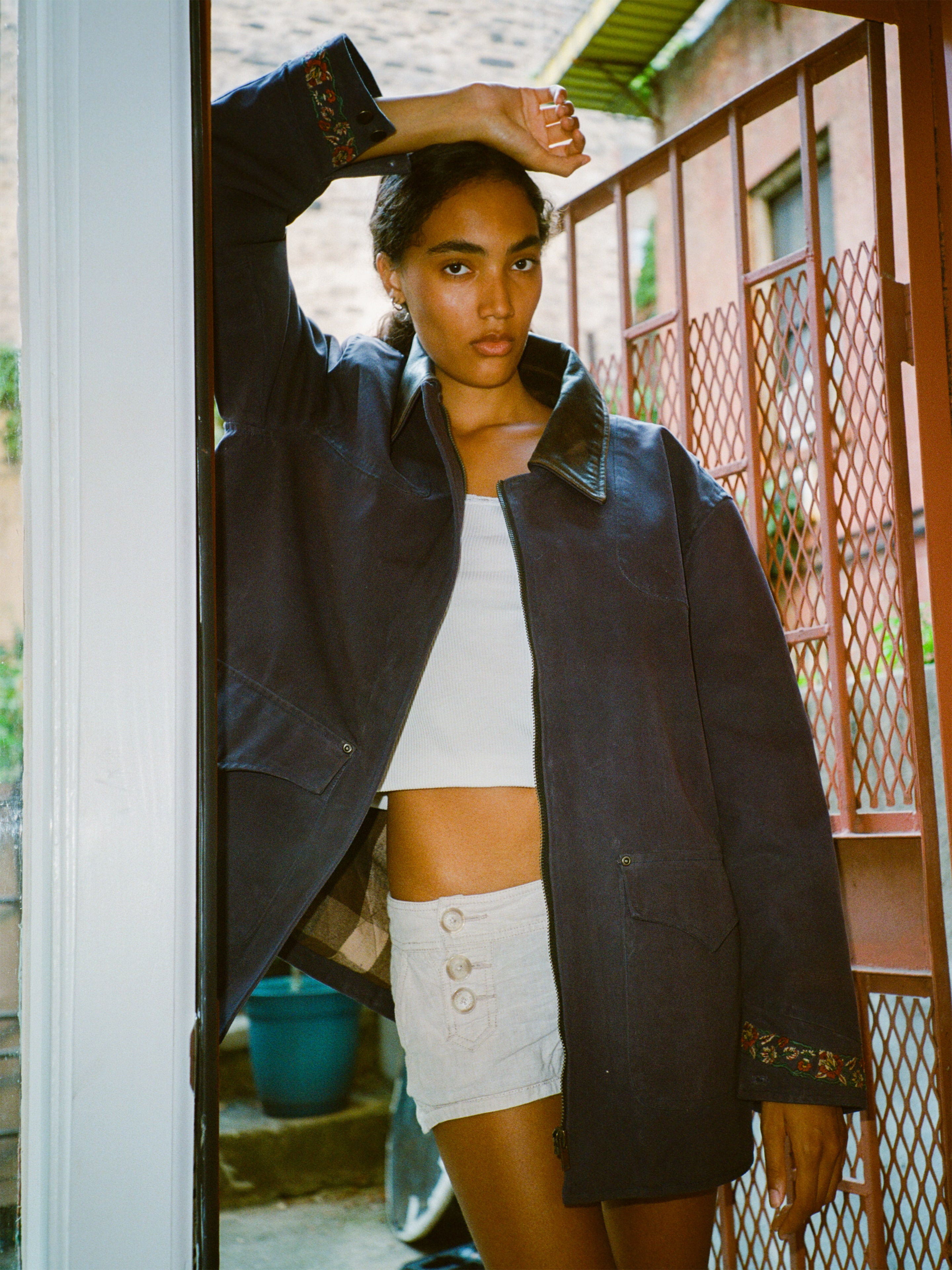 A woman poses in a doorway by a metal gate, one arm raised, wearing the Found Canvas Distressed Barn Jacket with a distressed leather collar over a white crop top and high-waisted shorts.