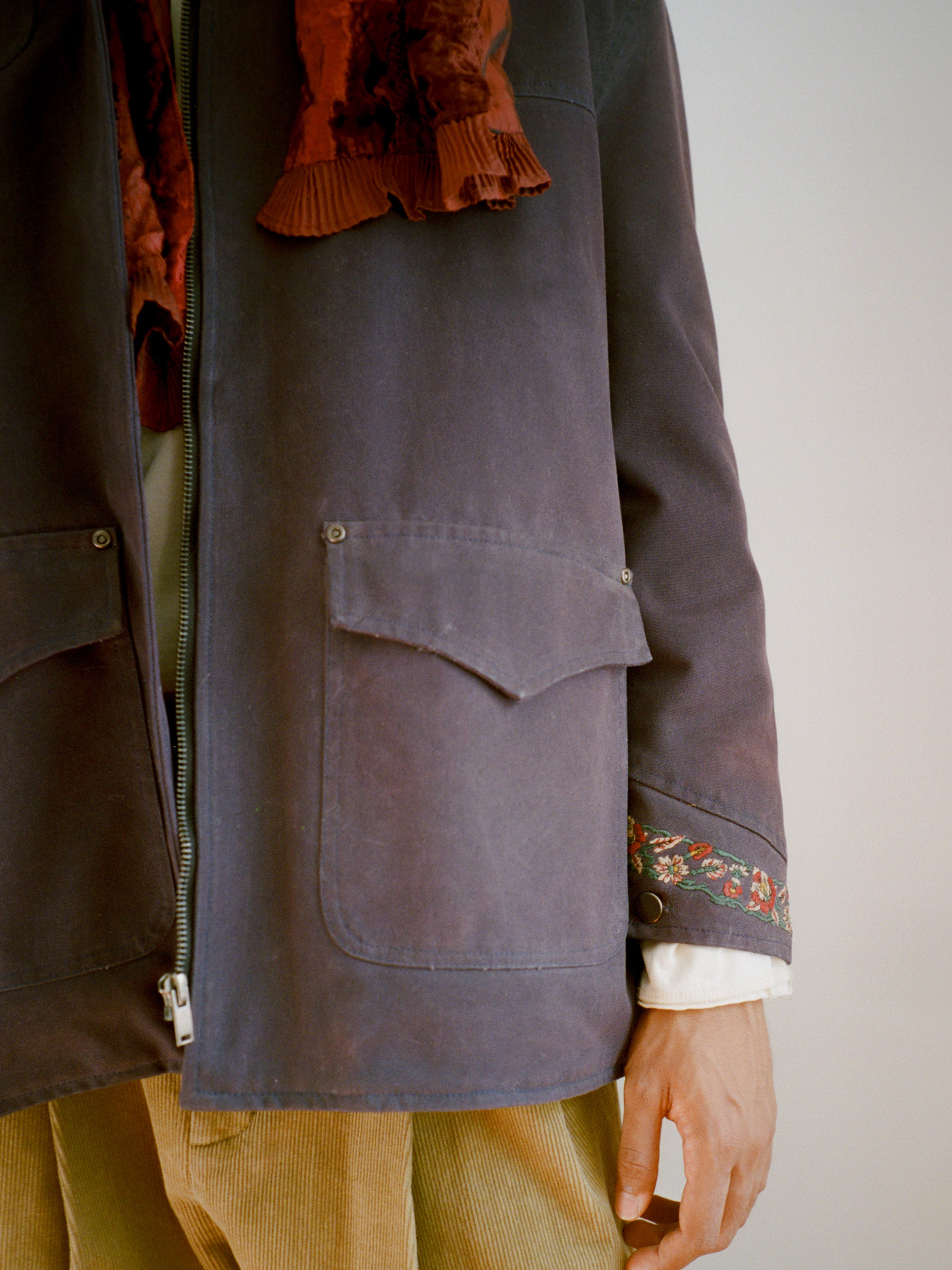 A person wears Found’s Canvas Distressed Barn Jacket—unisex, navy, relaxed fit with large pockets and a distressed leather collar—styled with a patterned scarf, white floral-embroidered sleeve, and beige corduroy pants.