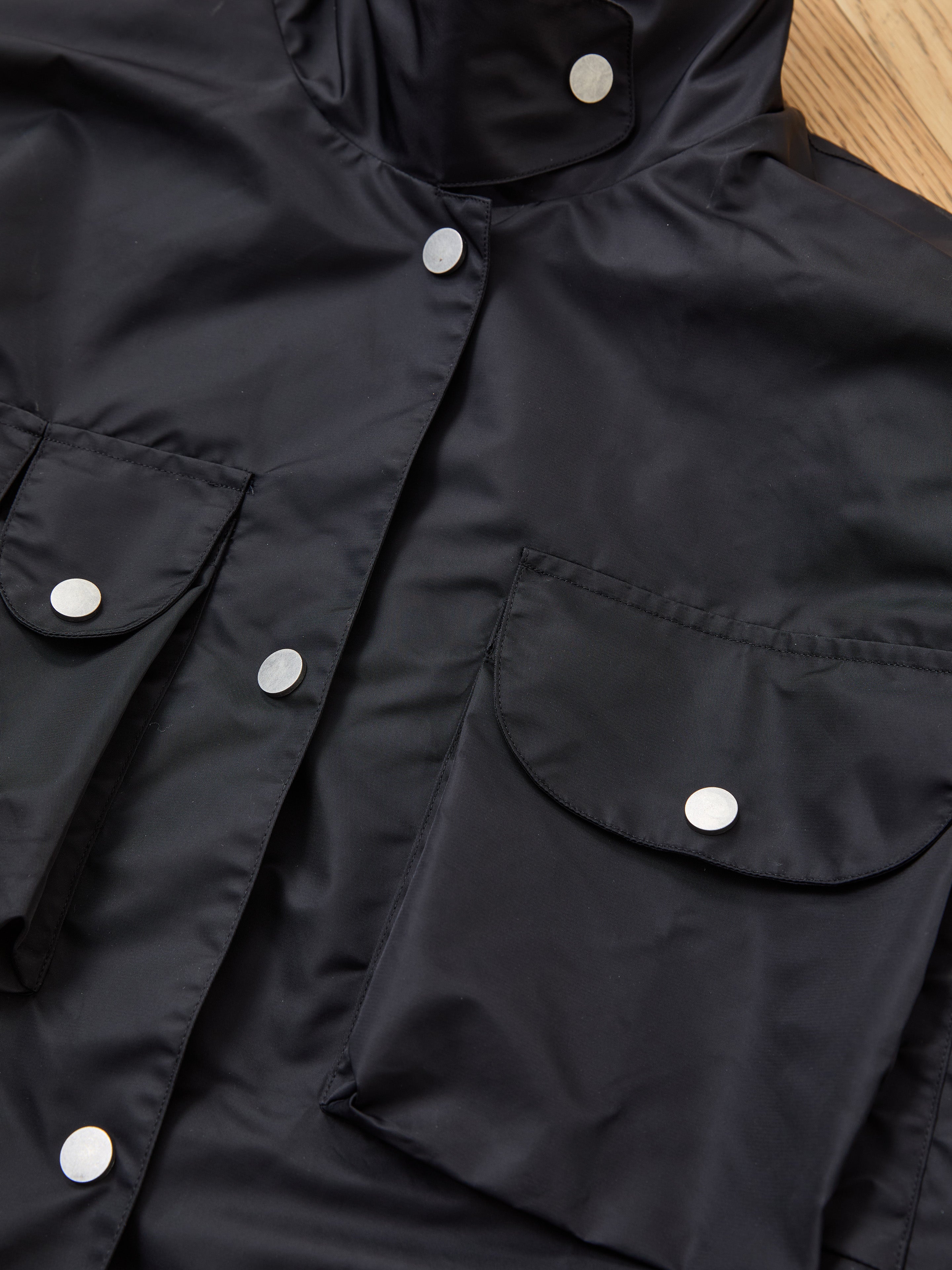 Close-up of the FOUND Cargo Nylon Jacket, a black unisex design with silver snap buttons and flap pockets, displayed on a wooden surface.