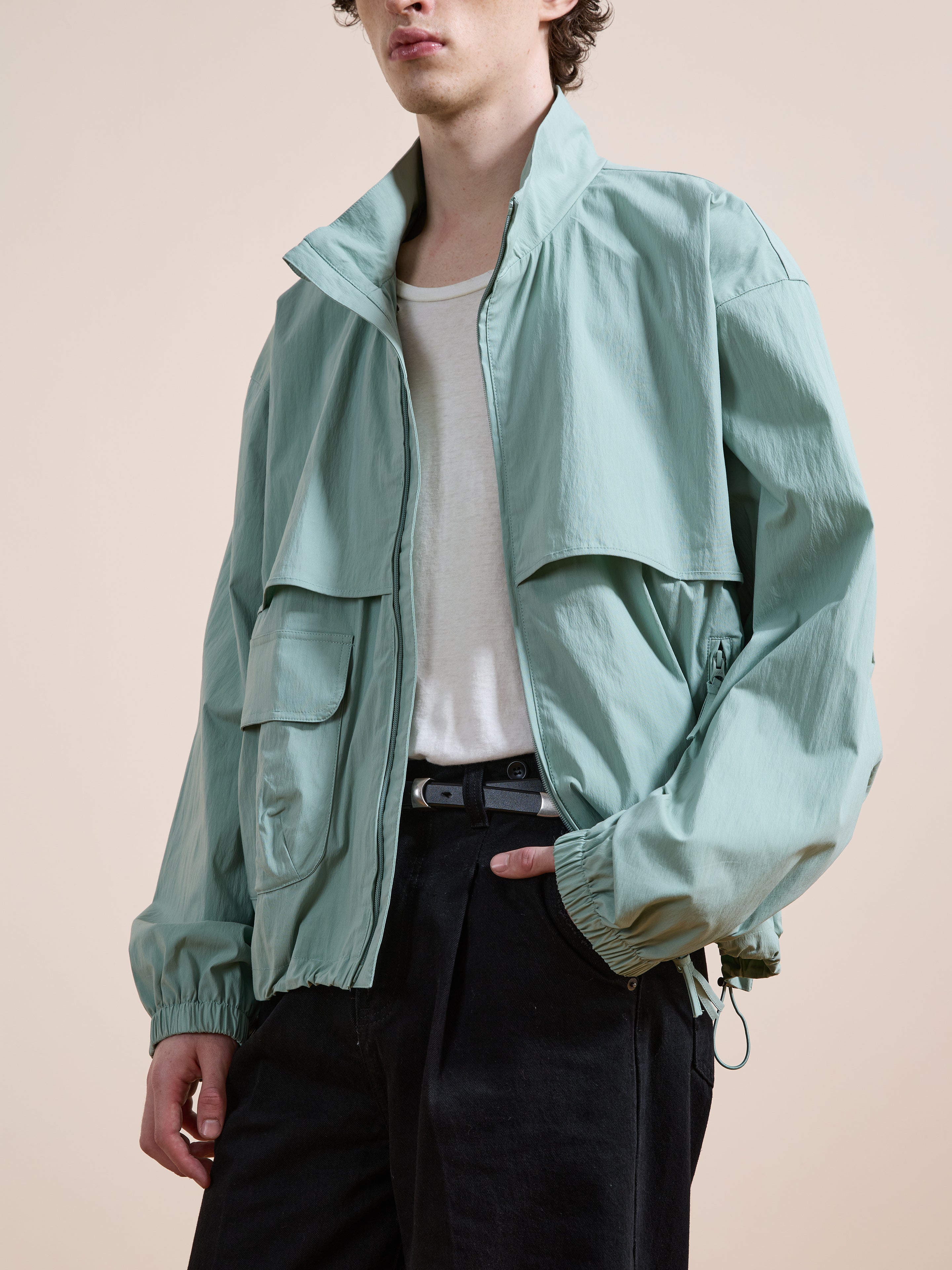 A person wears the FOUND Nylon Pocket Track Jacket in light green over a white t-shirt and black pants, standing against a plain background.