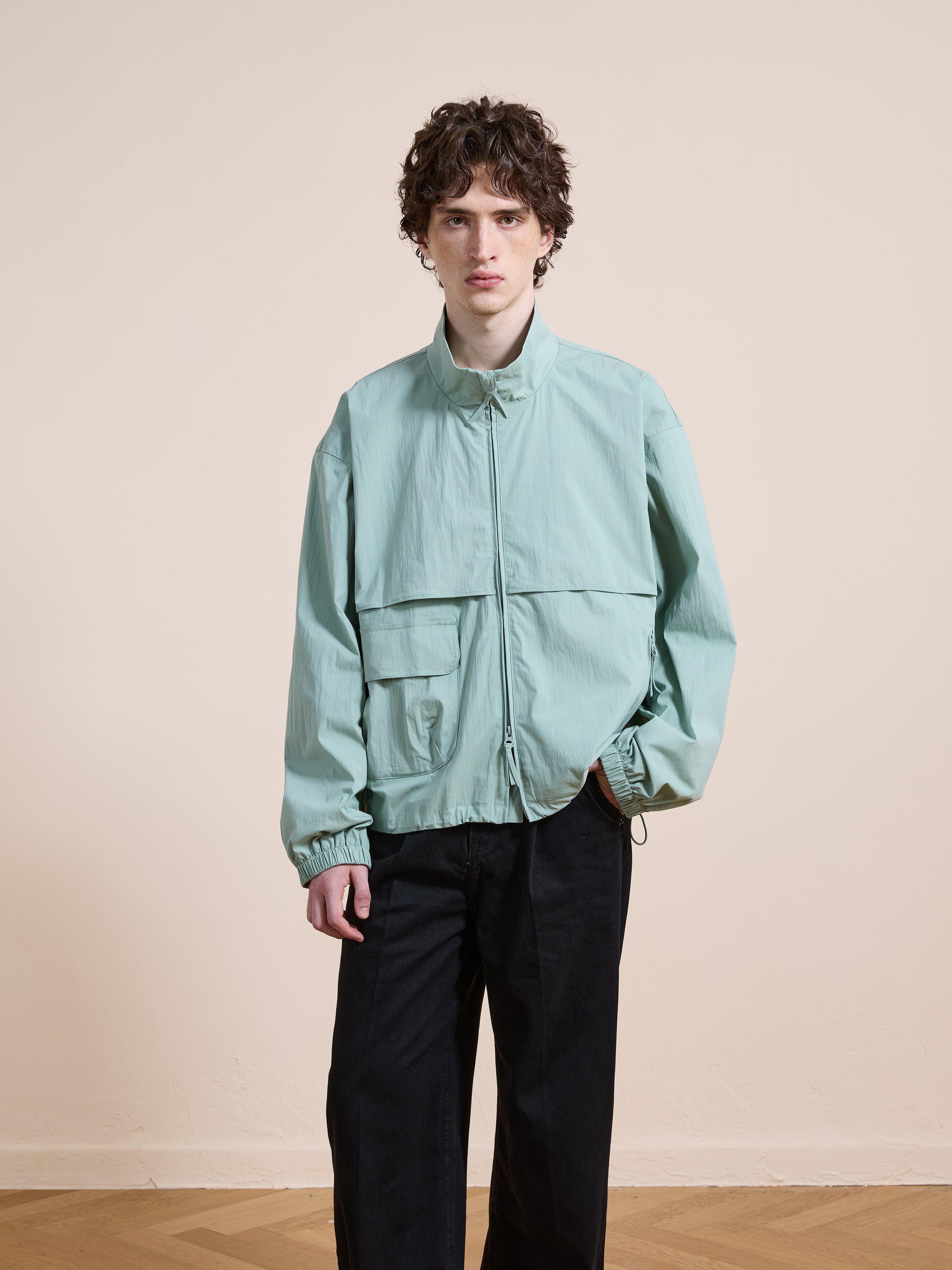 A person with curly hair wears the FOUND Nylon Pocket Track Jacket in light green, styled with black pants, standing indoors against a plain beige wall.