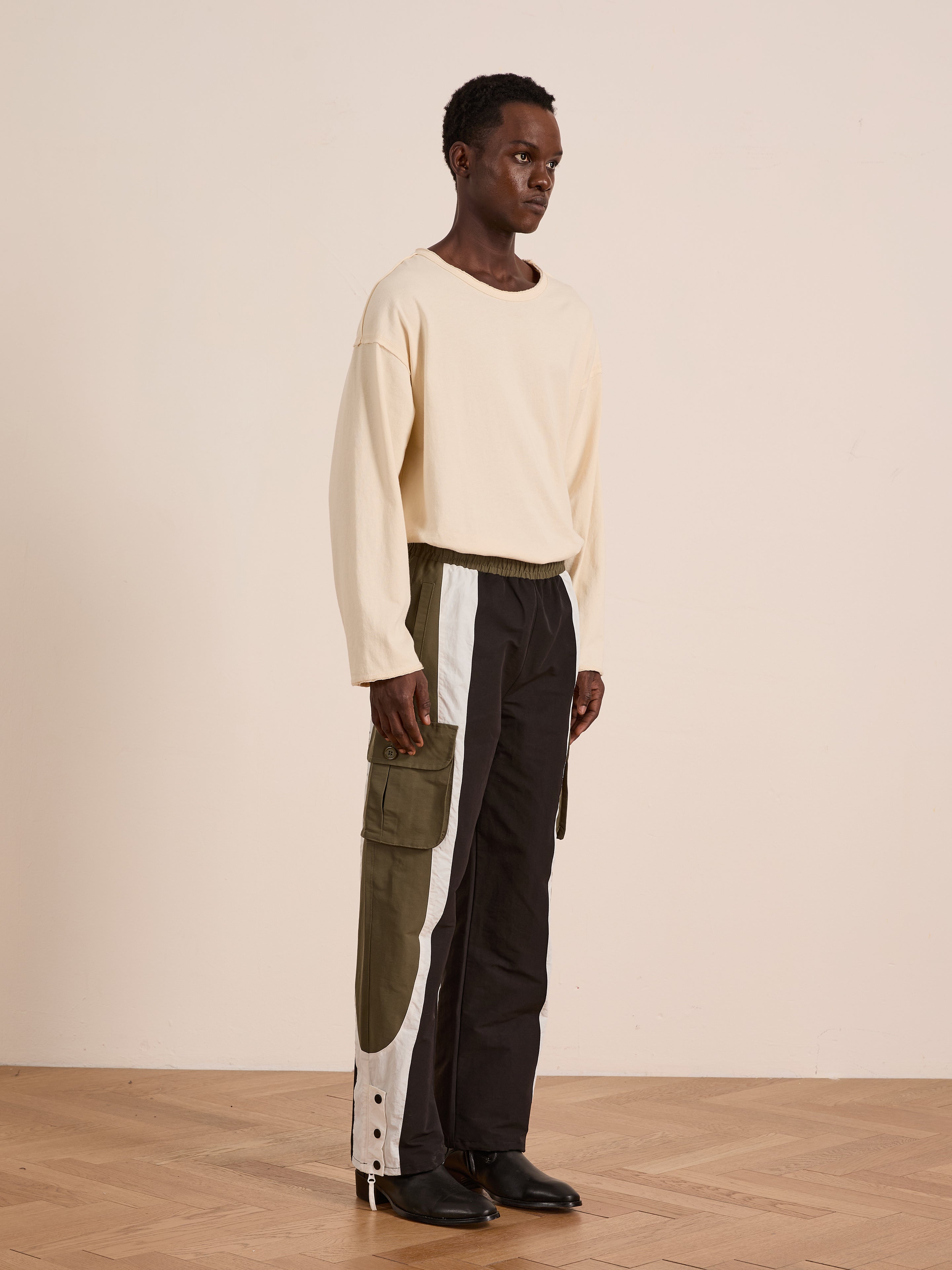 Indoors, a person wears a cream long-sleeve shirt, black shoes, and Found Sporting Club Track Pants—unisex sportswear featuring green and white panels and an adjustable waistband—standing on a wooden floor.