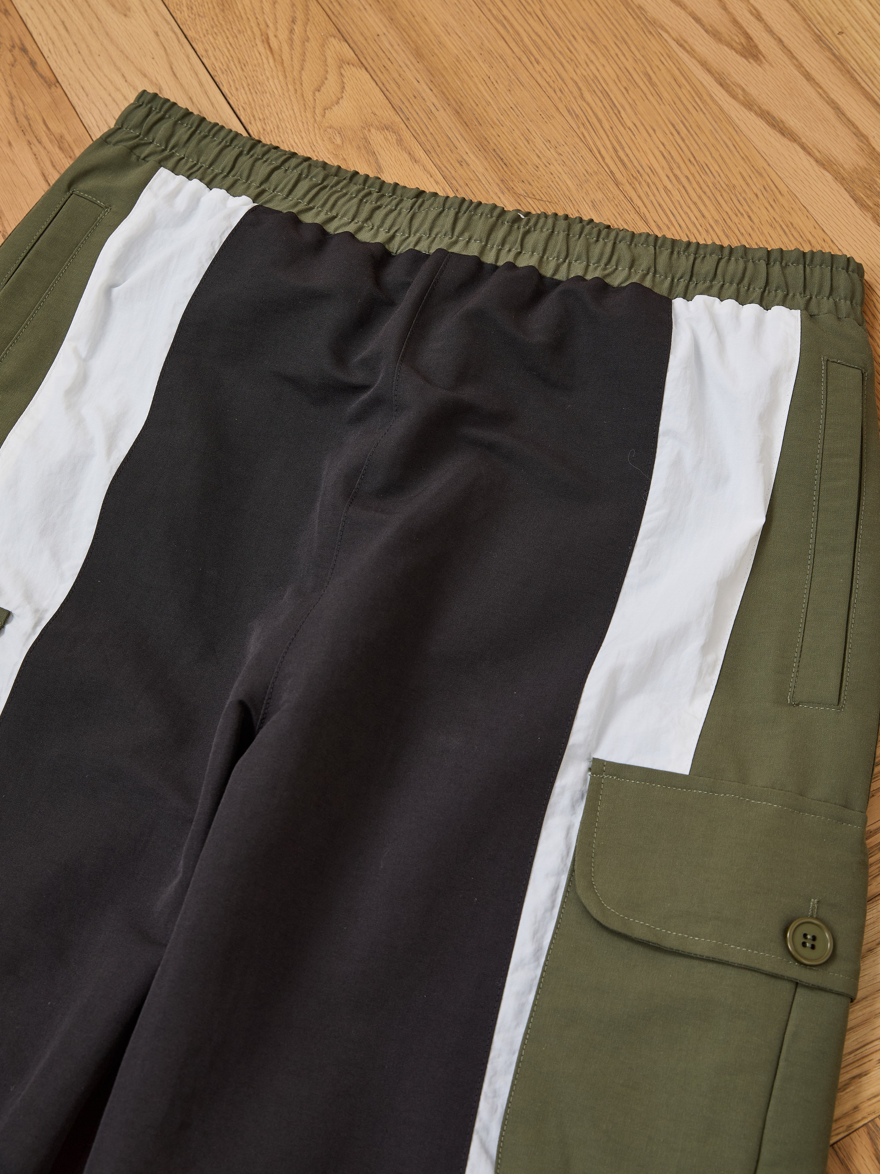 Found Sporting Club Track Pants—unisex, color-blocked in green, black, and white—with an adjustable waistband and side button-flap pocket, shown on a wooden floor.