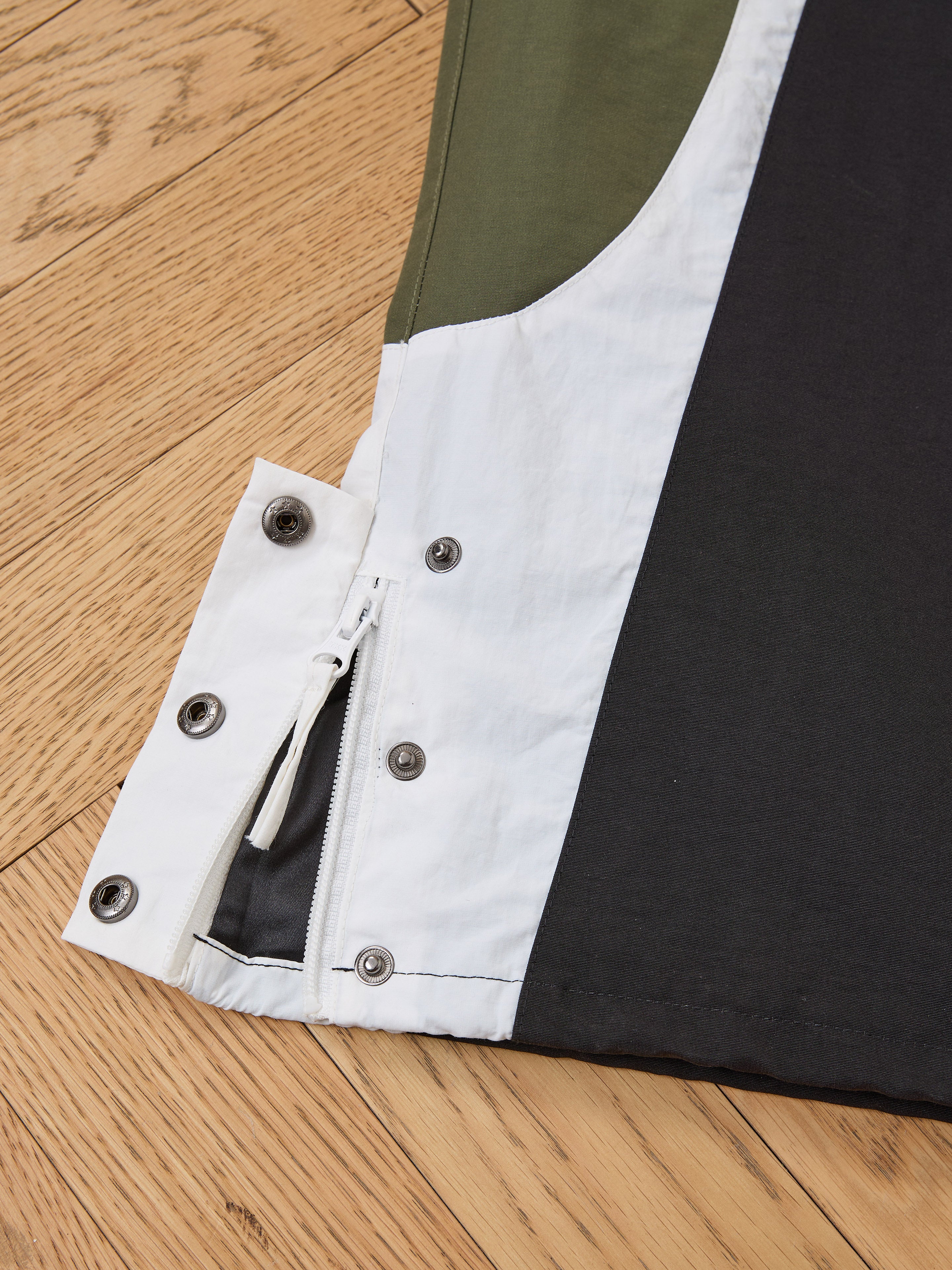 Close-up of Found Sporting Club Track Pants hem, showing white, black, and green color blocks with a zipper, metal snap buttons, adjustable waistband, and stitching details on a wooden floor.