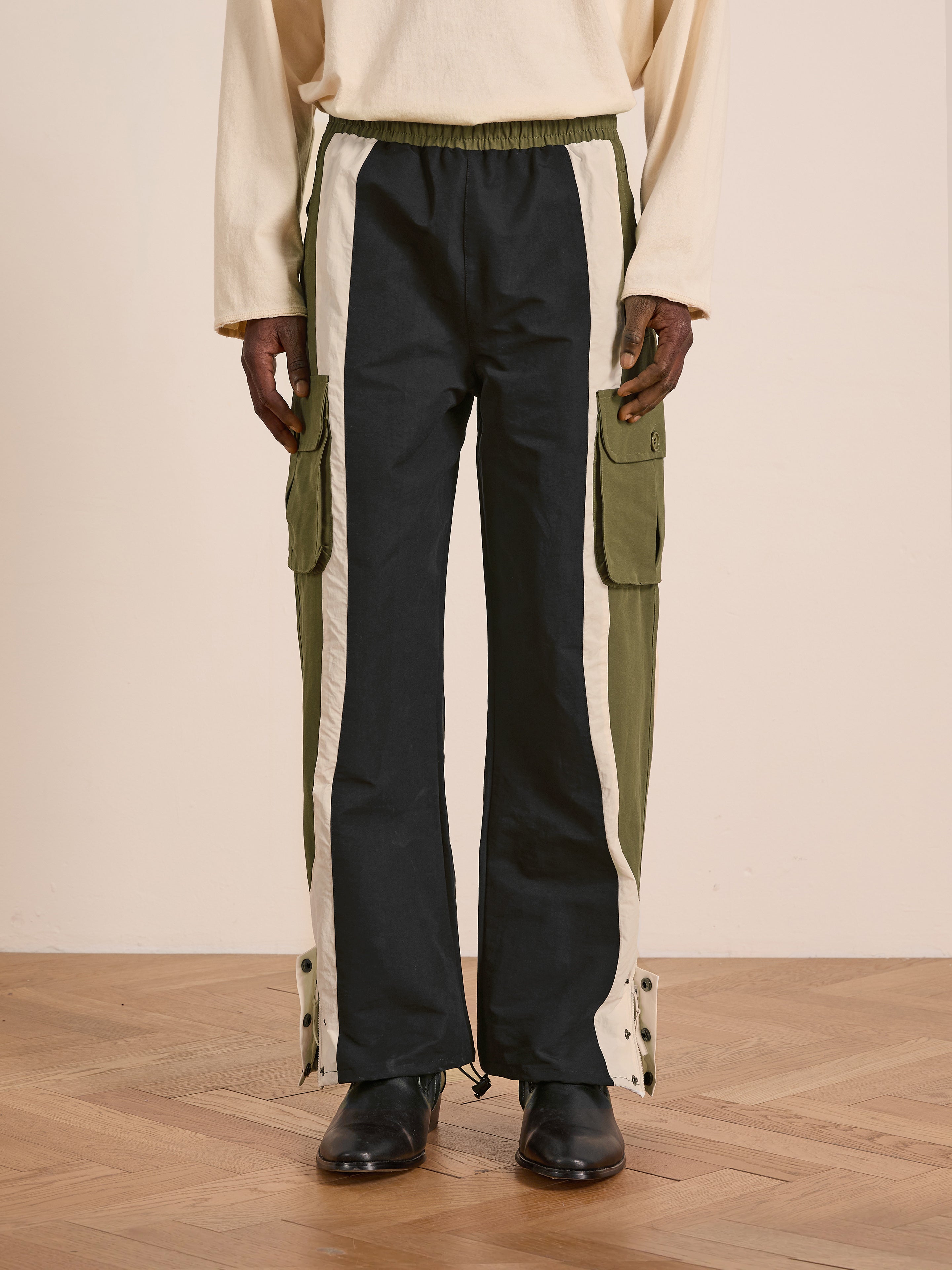 A person wears Found Sporting Club Track Pants—relaxed fit colorblock cargos with black, olive green, and white panels—paired with a cream long-sleeve shirt and black shoes, standing on a wooden floor.