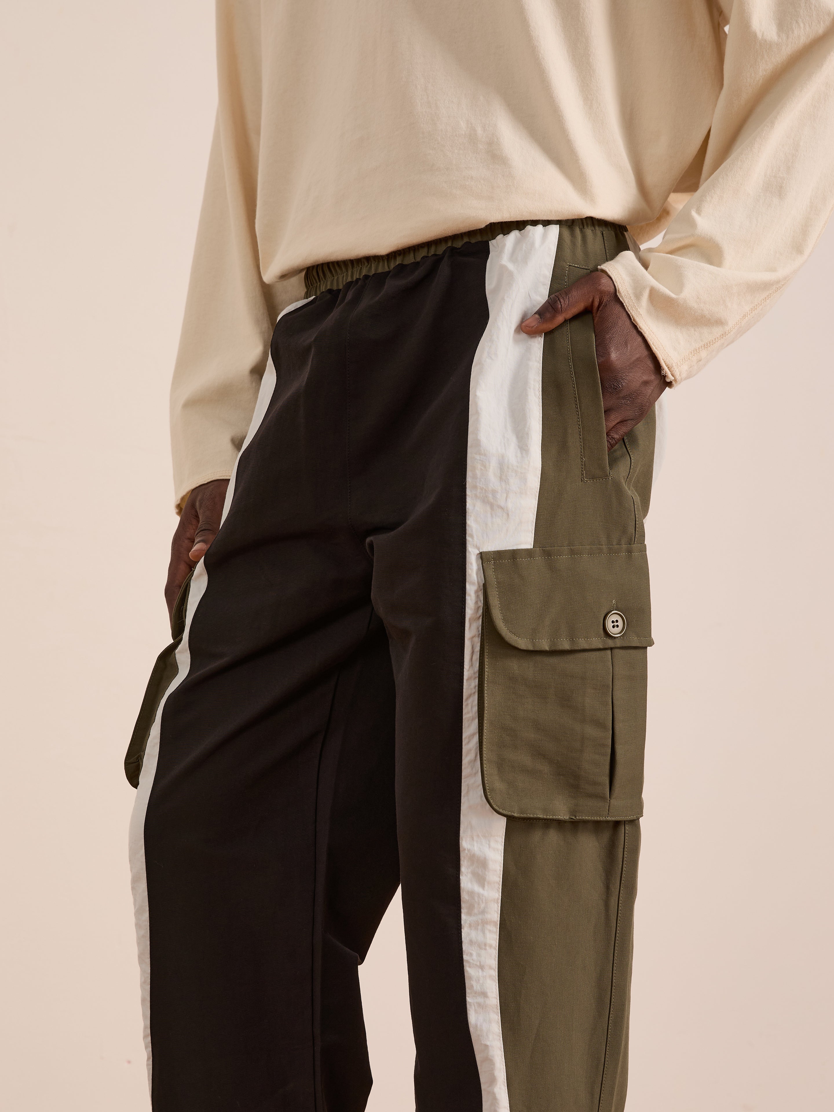 A person wearing Found's Sporting Club Track Pants—unisex color-block pants with black, white, and olive green panels, large side pockets, and an adjustable waistband—paired with a beige long-sleeve shirt.