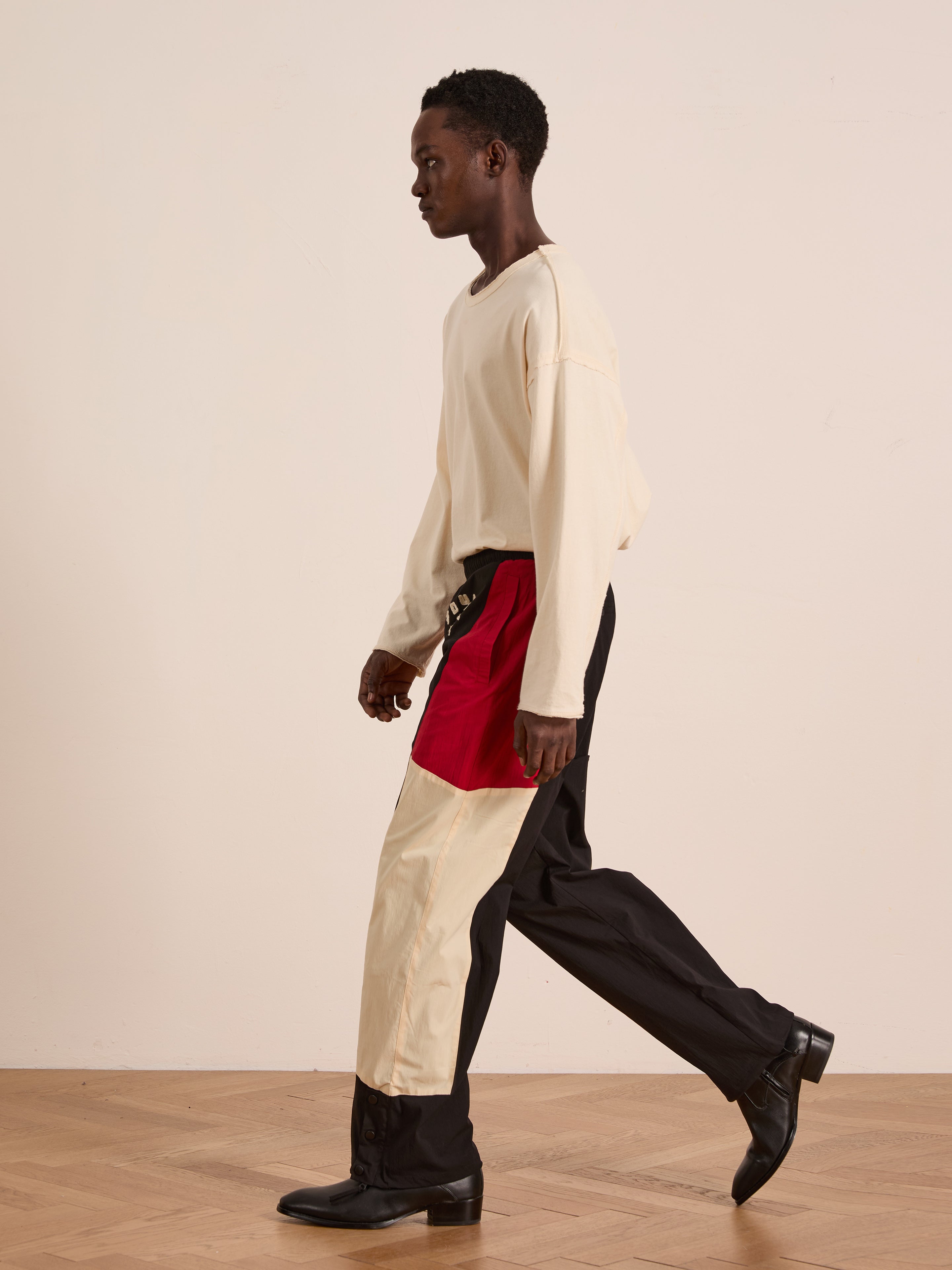 A person walks indoors in a cream long-sleeve shirt, black shoes, and Found’s Pine and Rose Track Pants featuring a unisex relaxed fit in red, cream, and black. The background is plain and minimal.