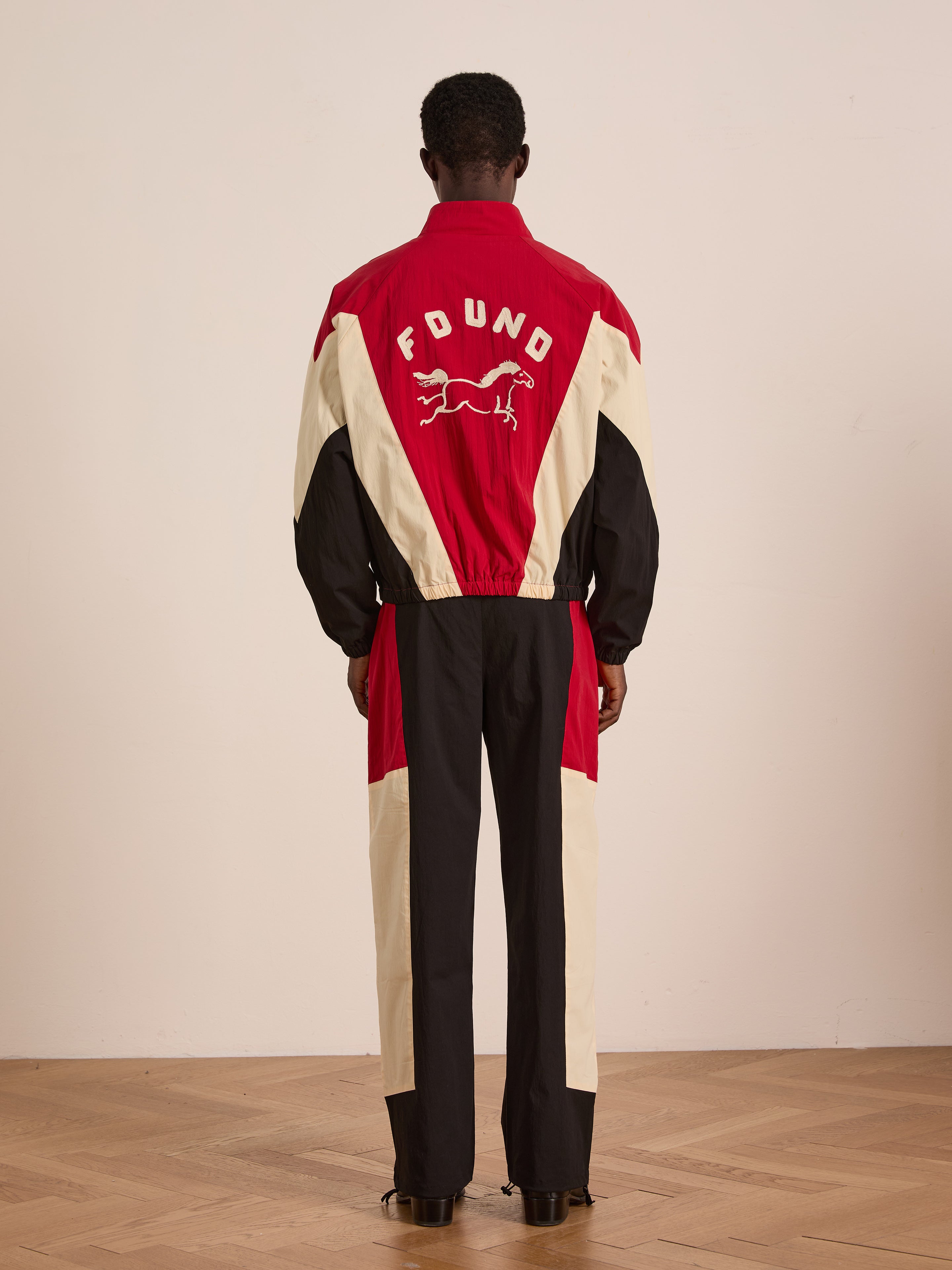 A person stands facing away, wearing Found’s Pine and Rose Track Pants—a unisex relaxed-fit, red, black, and cream color-blocked tracksuit with "FOUND" and a running dog graphic on the back—paired with athletic nylon pants.