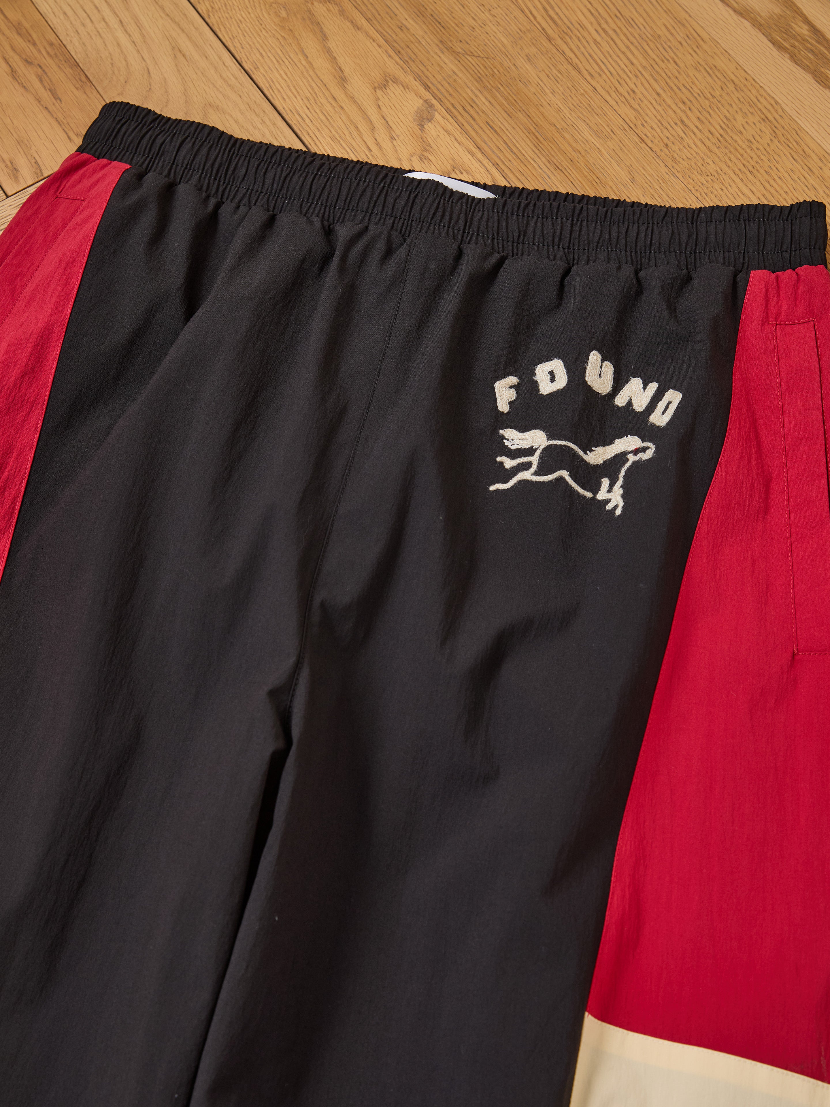 Pine and Rose Track Pants by Found with a unisex relaxed fit, elastic waistband, "FOUND" text, and running dog graphic on the left thigh, shown laid flat on a wooden floor.