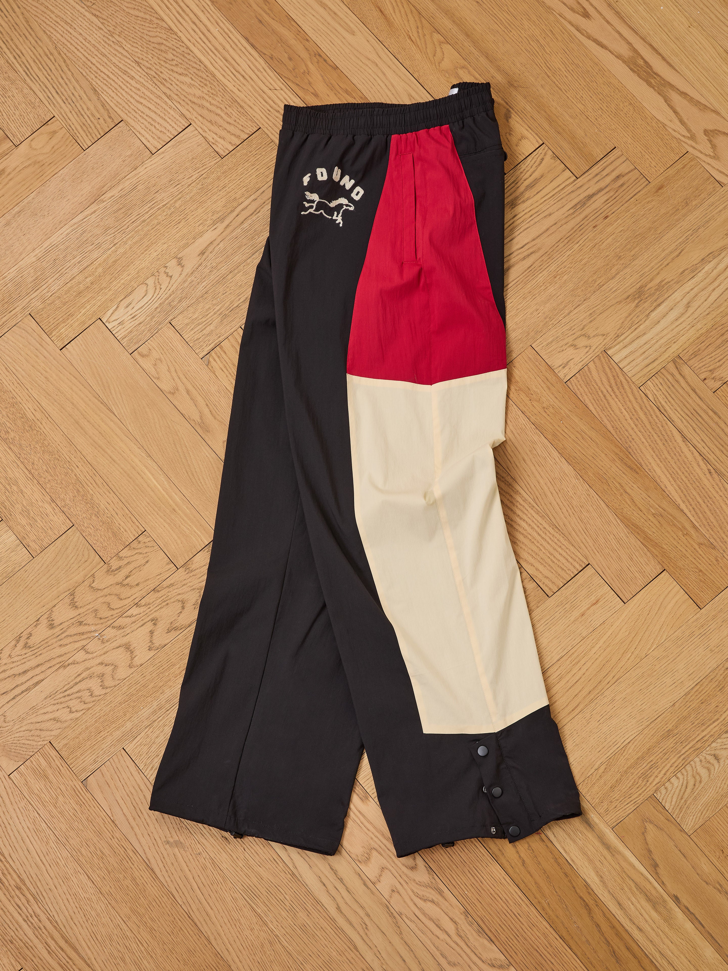 Pine and Rose Track Pants by Found in black, red, and beige with "YOUNG" text and graphic on the waist, unisex relaxed fit, displayed flat on a wooden herringbone floor.