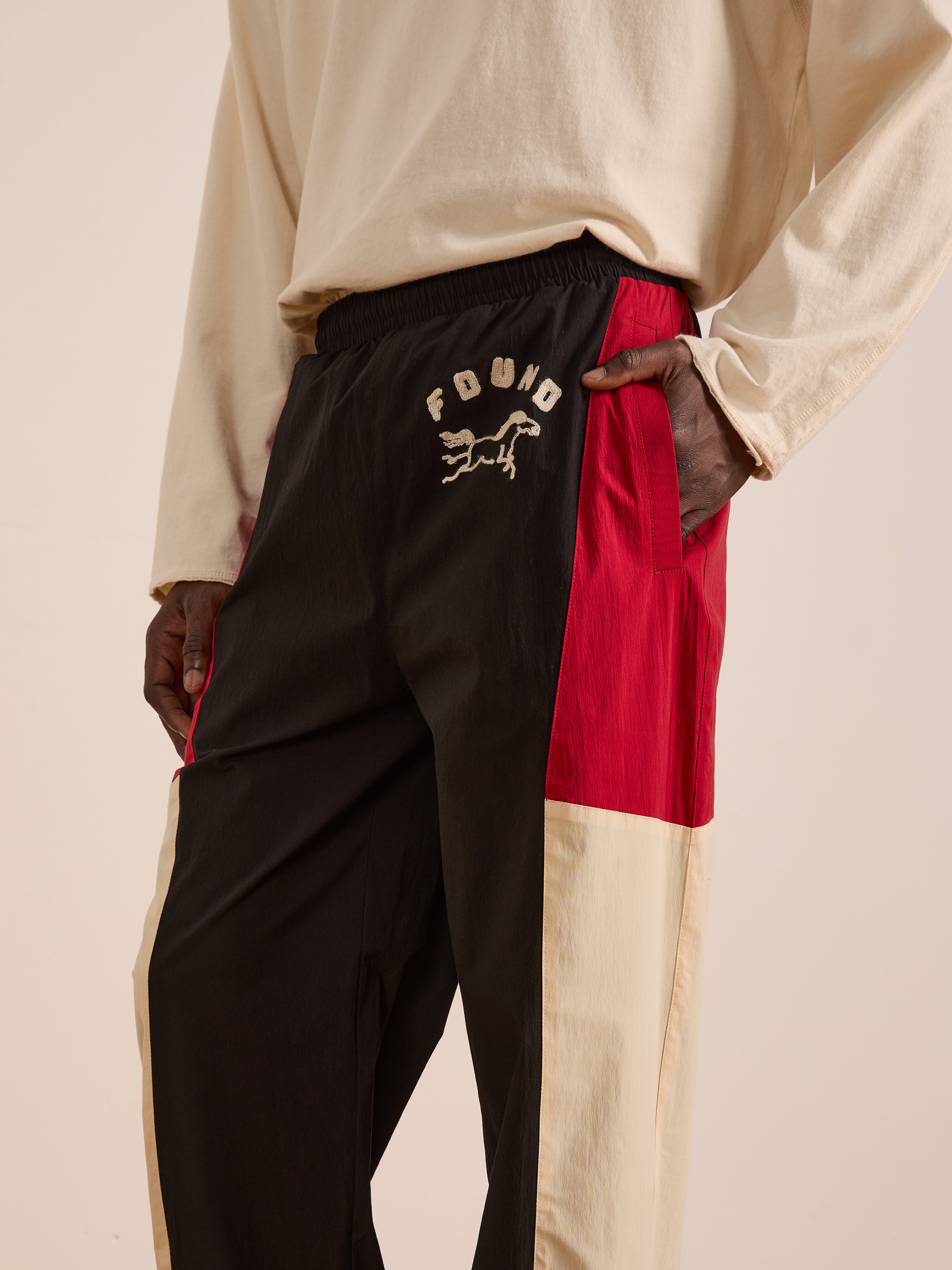A person wears Found Pine and Rose Track Pants in black, red, and beige with a "FOUND" graphic on the left thigh, paired with a beige long-sleeve shirt. These unisex nylon pants feature a relaxed fit for sporty style.