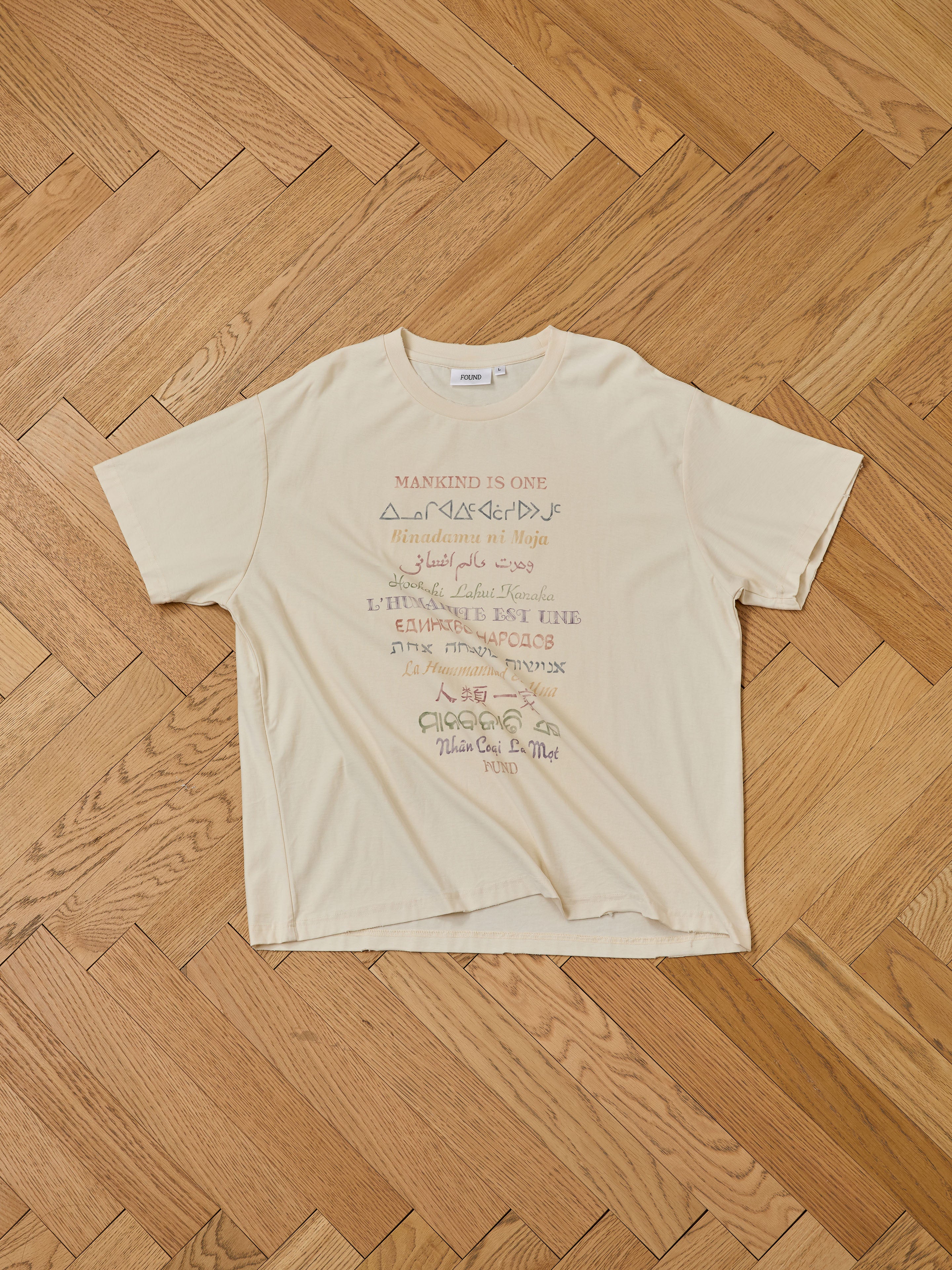 The FOUND One Humanity Tee is a beige unisex, relaxed fit short-sleeve t-shirt featuring multicolored text and script designs on the front, displayed flat on a wooden herringbone floor.