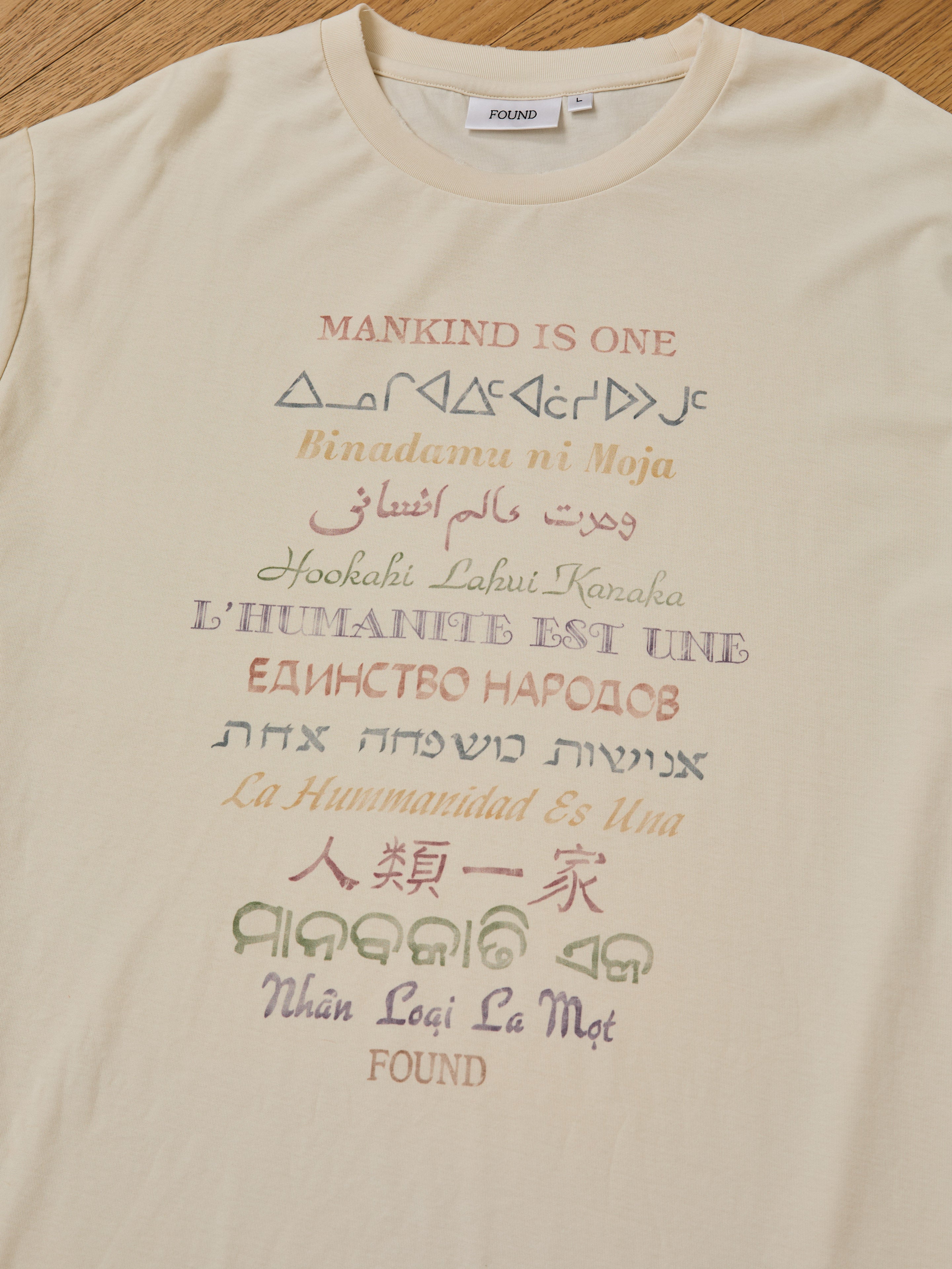 The One Humanity Tee by FOUND is a beige unisex vintage cotton shirt with a relaxed fit, featuring “Mankind is one” in many languages and scripts, plus FOUND branding near the collar and hem—express unity in style.