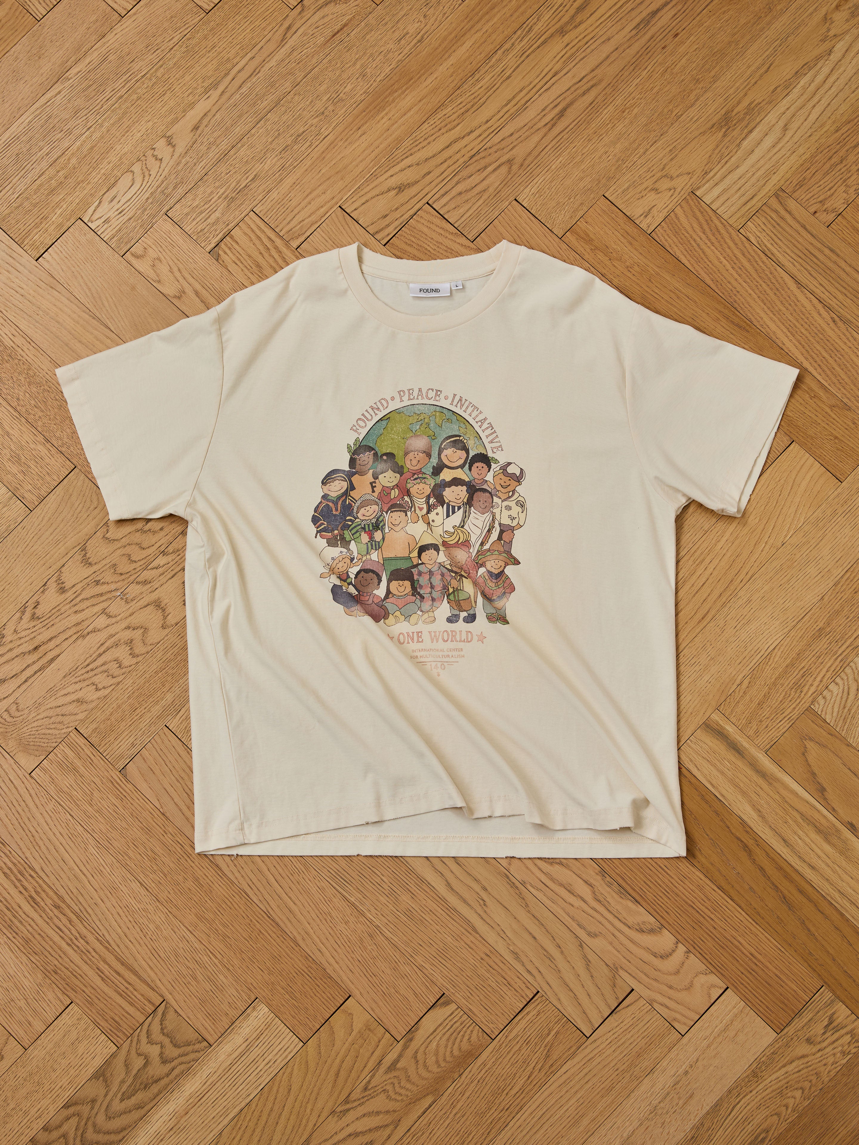 The One World Family Tee by FOUND is a cream-colored t-shirt featuring diverse children holding hands in a circle, with "KINDNESS • PEACE • UNITY" above and "ONE WORLD" below.