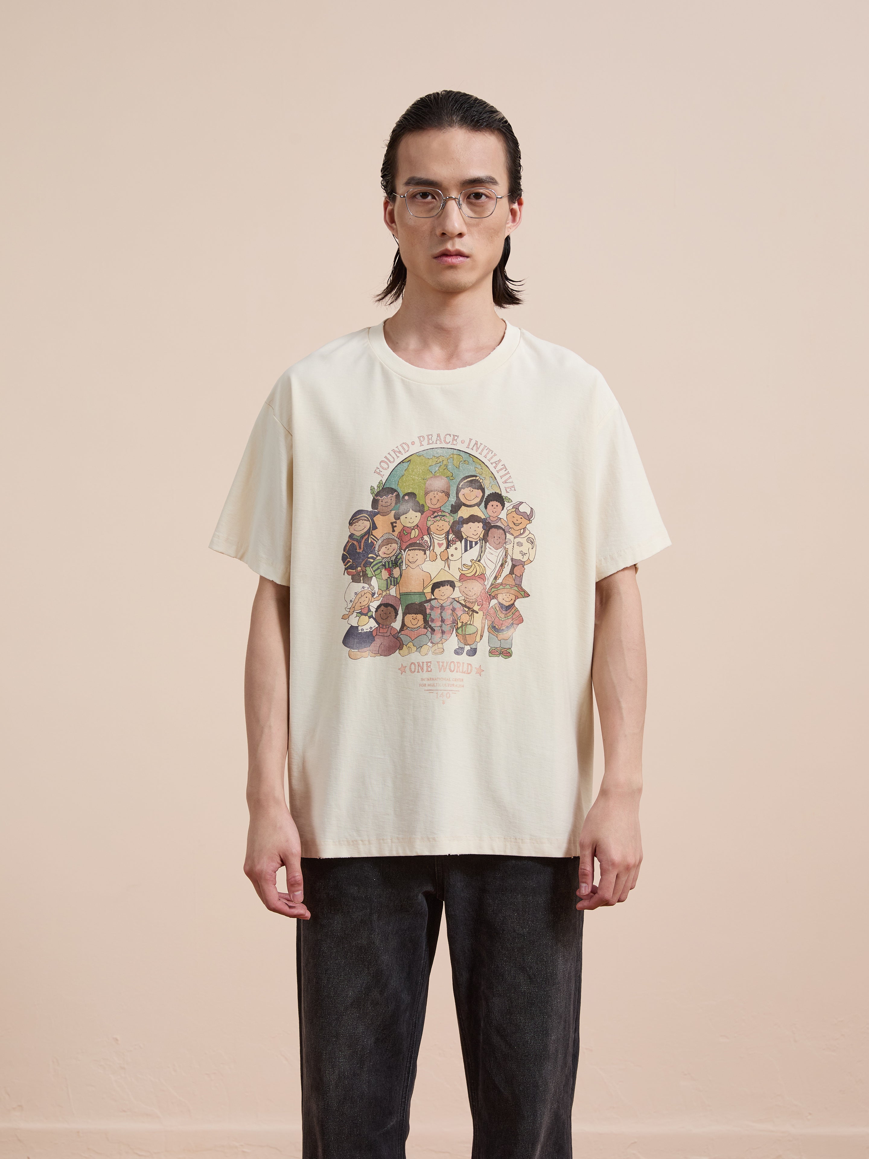 A person wearing glasses models the FOUND One World Family Tee, a cream-colored shirt with diverse faces and "One World" text, against a plain background.