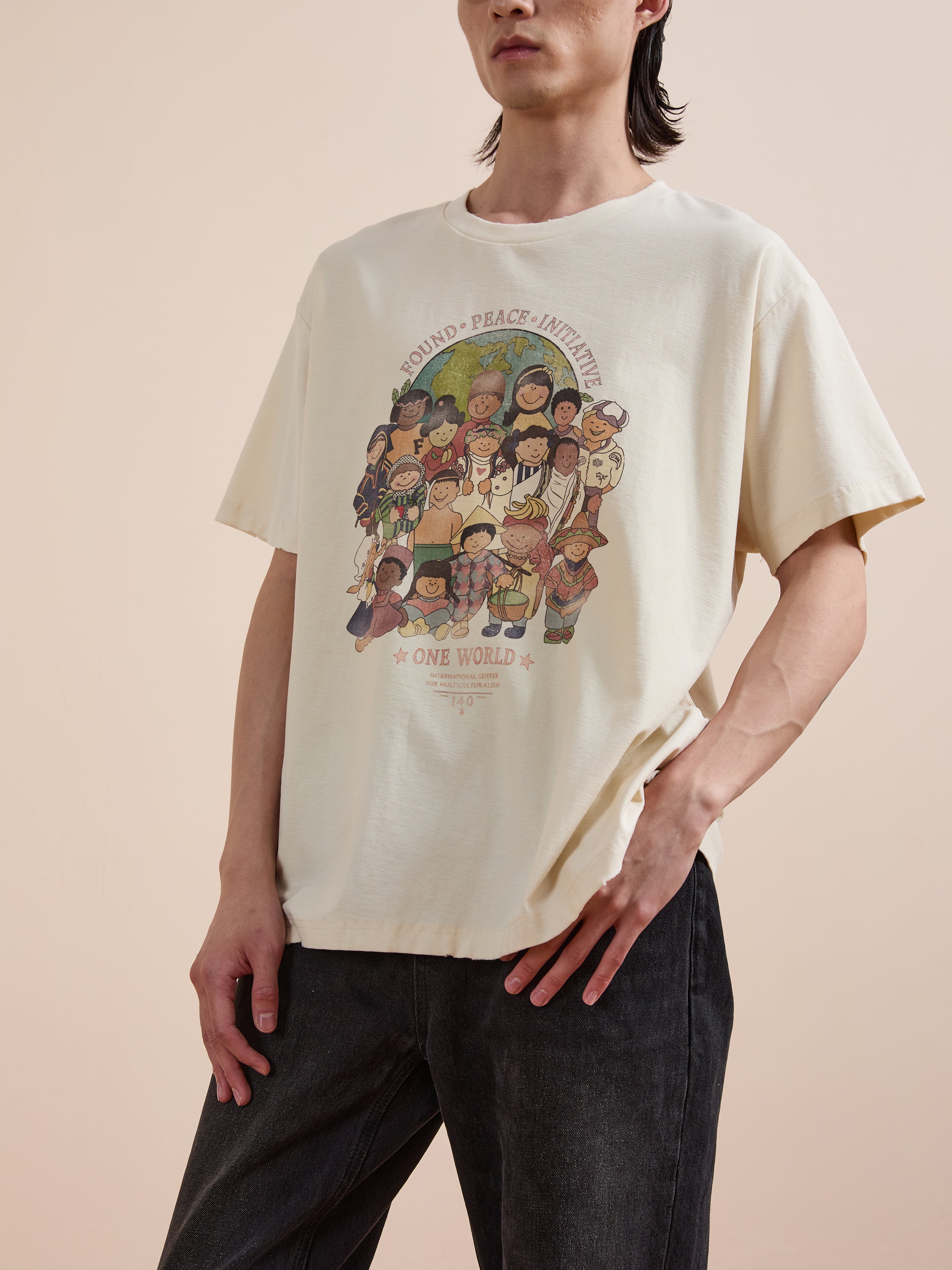 Wearing the FOUND One World Family Tee, a light-toned multicultural shirt featuring cartoon children and “Sound Peace Initiative One World,” a person stands against a neutral background.