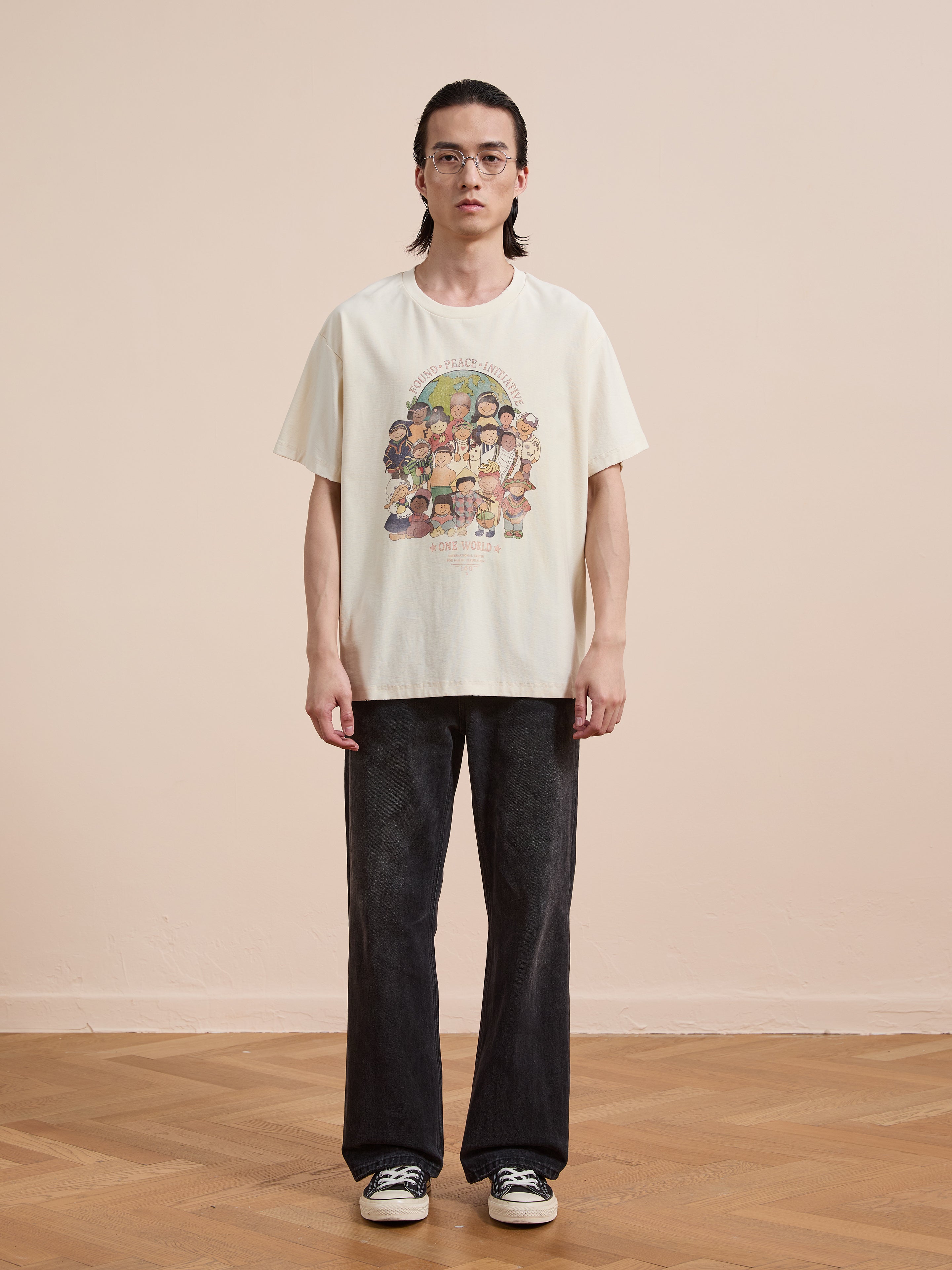 A person with long hair, glasses, and a neutral expression stands indoors on a wooden floor, wearing the FOUND One World Family Tee—paired with black jeans and black-and-white sneakers.