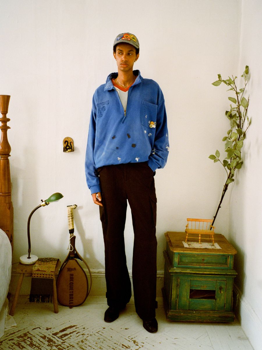 In a cozy room with a bed, guitar-like instrument, green nightstand, plant, and lamp, a person sports FOUND's Painters Pullover Sweatshirt with utilitarian flair along with dark pants and a cap.