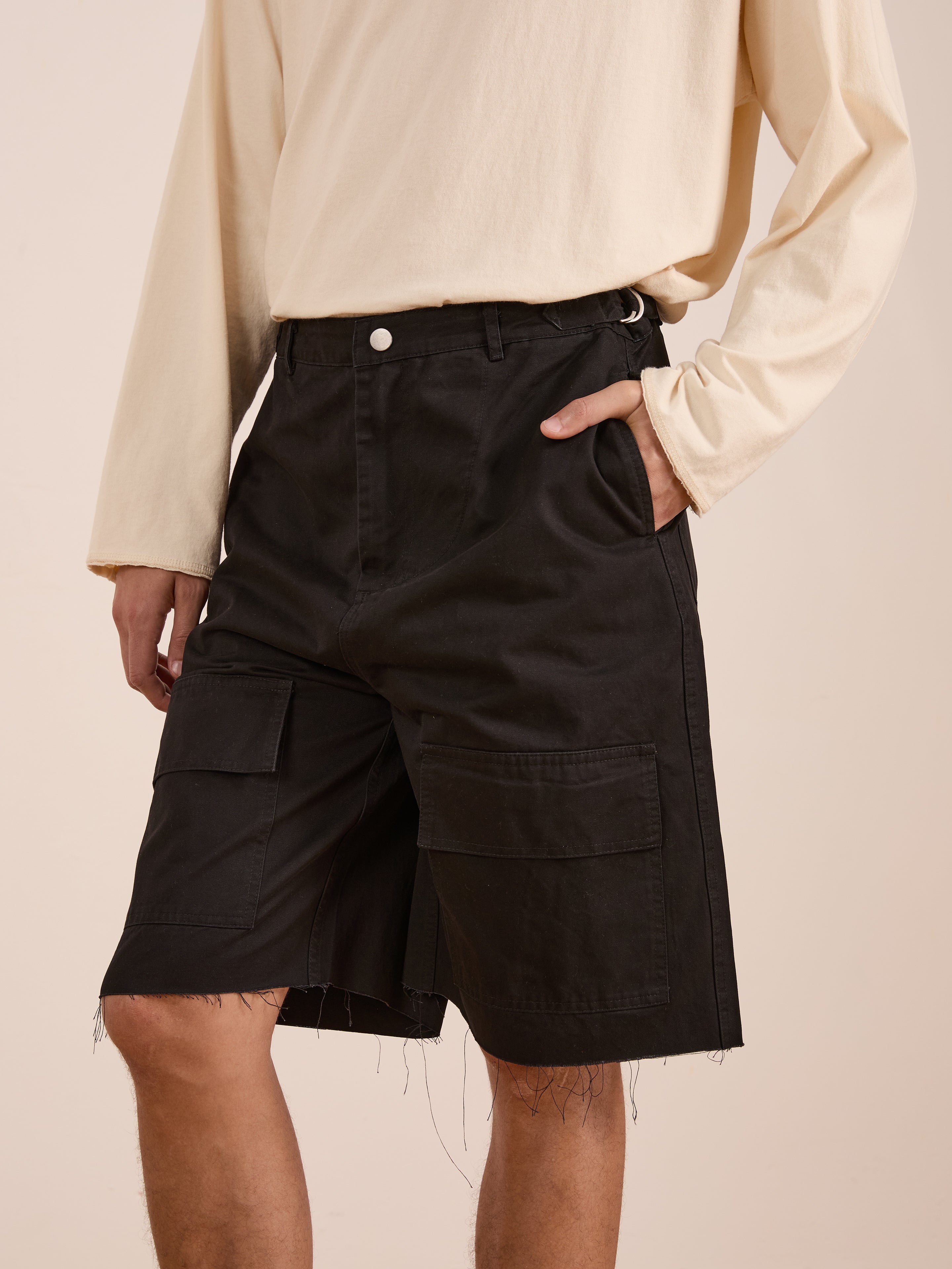 A person wearing a cream long-sleeve shirt and black Found Parachute Cargo Twill Shorts with frayed hems, standing with one hand in their pocket.