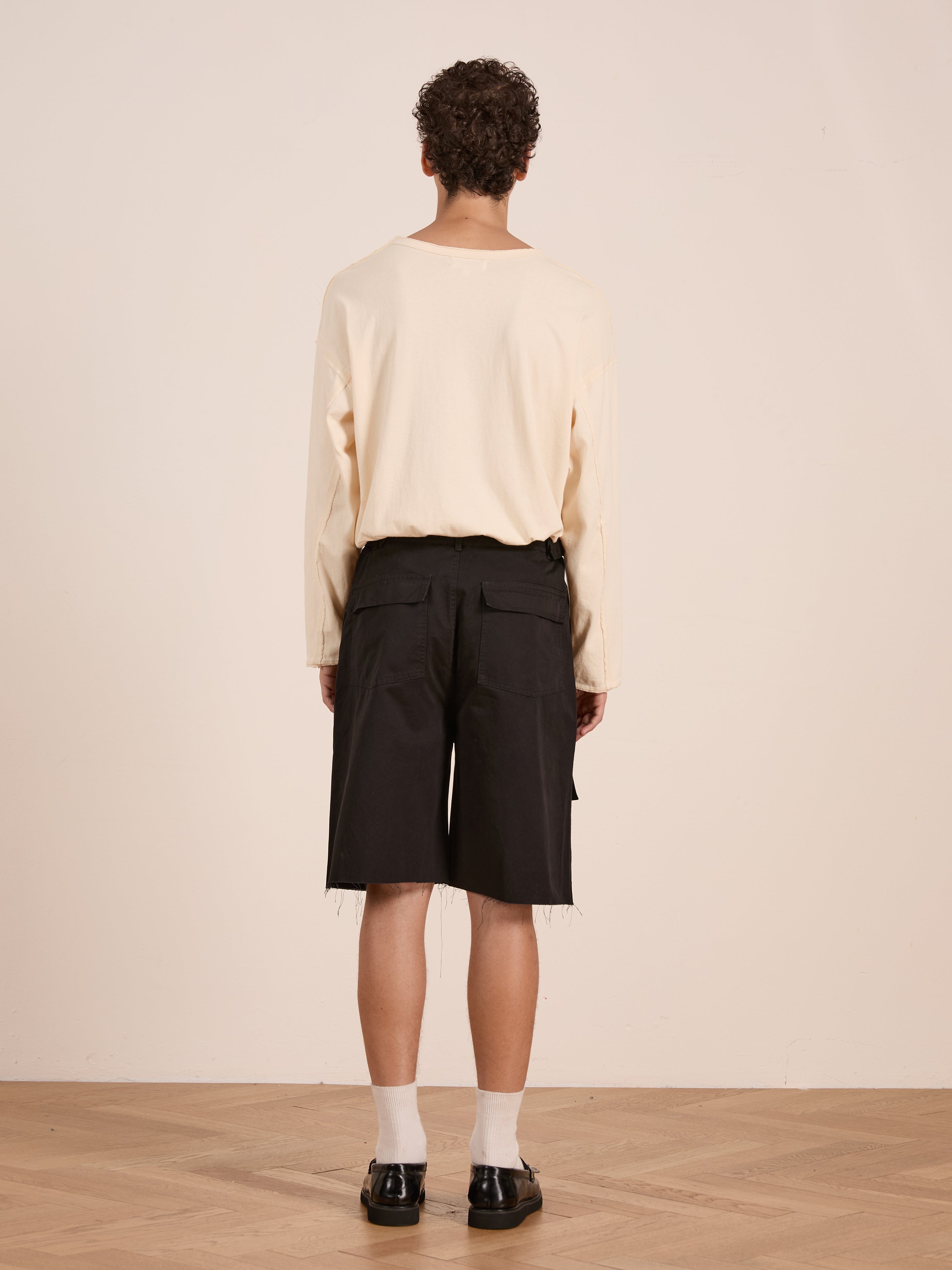 A person stands indoors facing away, wearing a long-sleeve beige shirt, black Found Parachute Cargo Twill Shorts, white socks, and black shoes on a wooden floor.