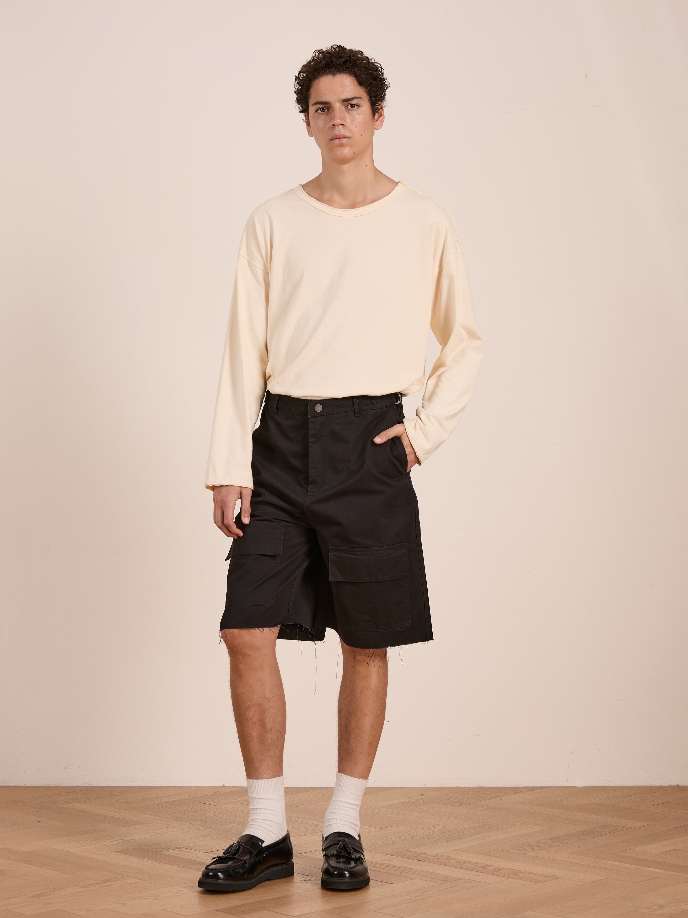Indoors on a wooden floor with a light backdrop, a person wears Found's Parachute Cargo Twill Shorts in black, paired with a cream long-sleeve shirt, white socks, and black loafers.