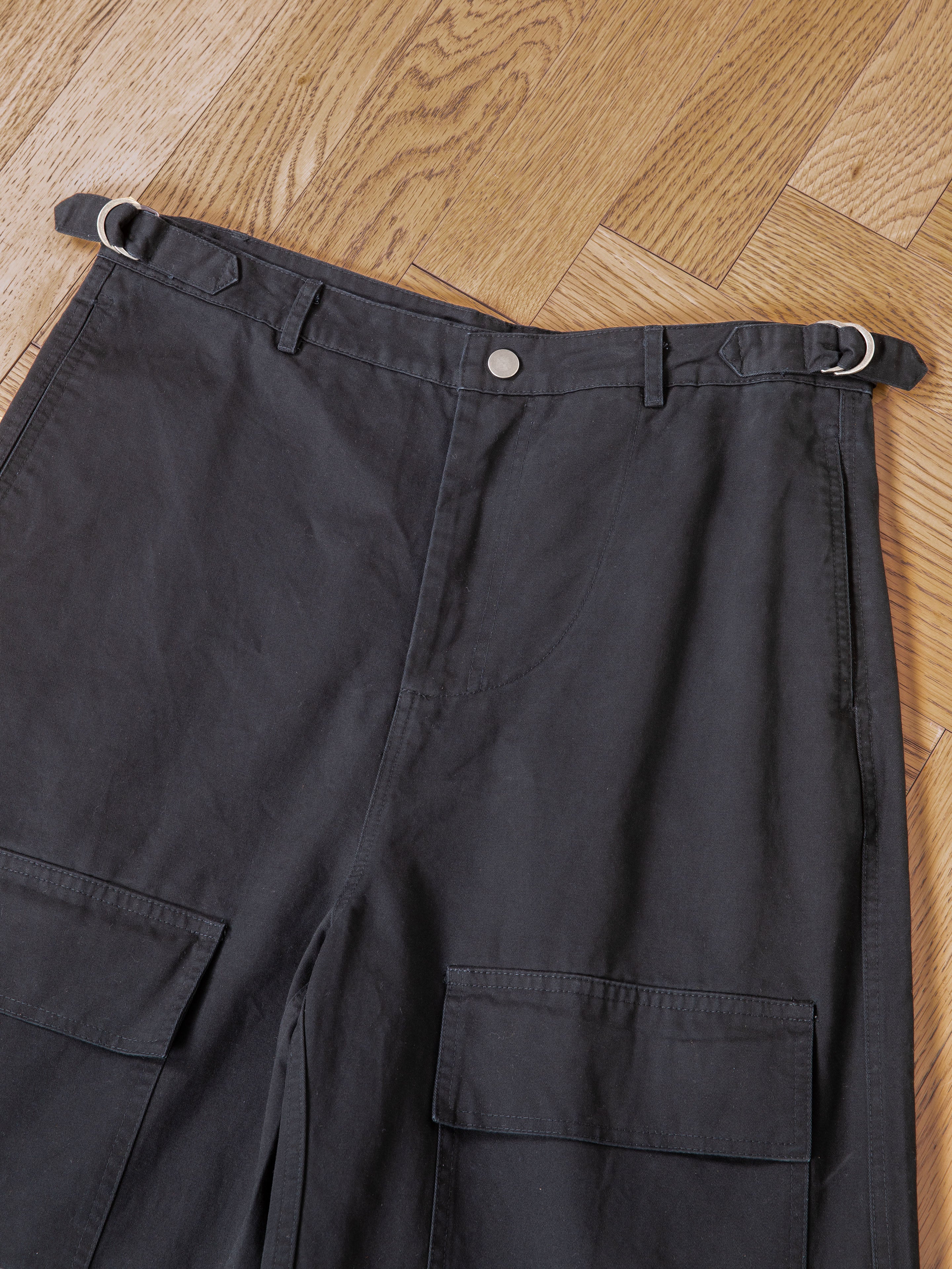 The Found Parachute Cargo Twill Shorts feature large front pockets and adjustable waist tabs in black, laid flat on a wooden floor—ideal for versatile utility wear.