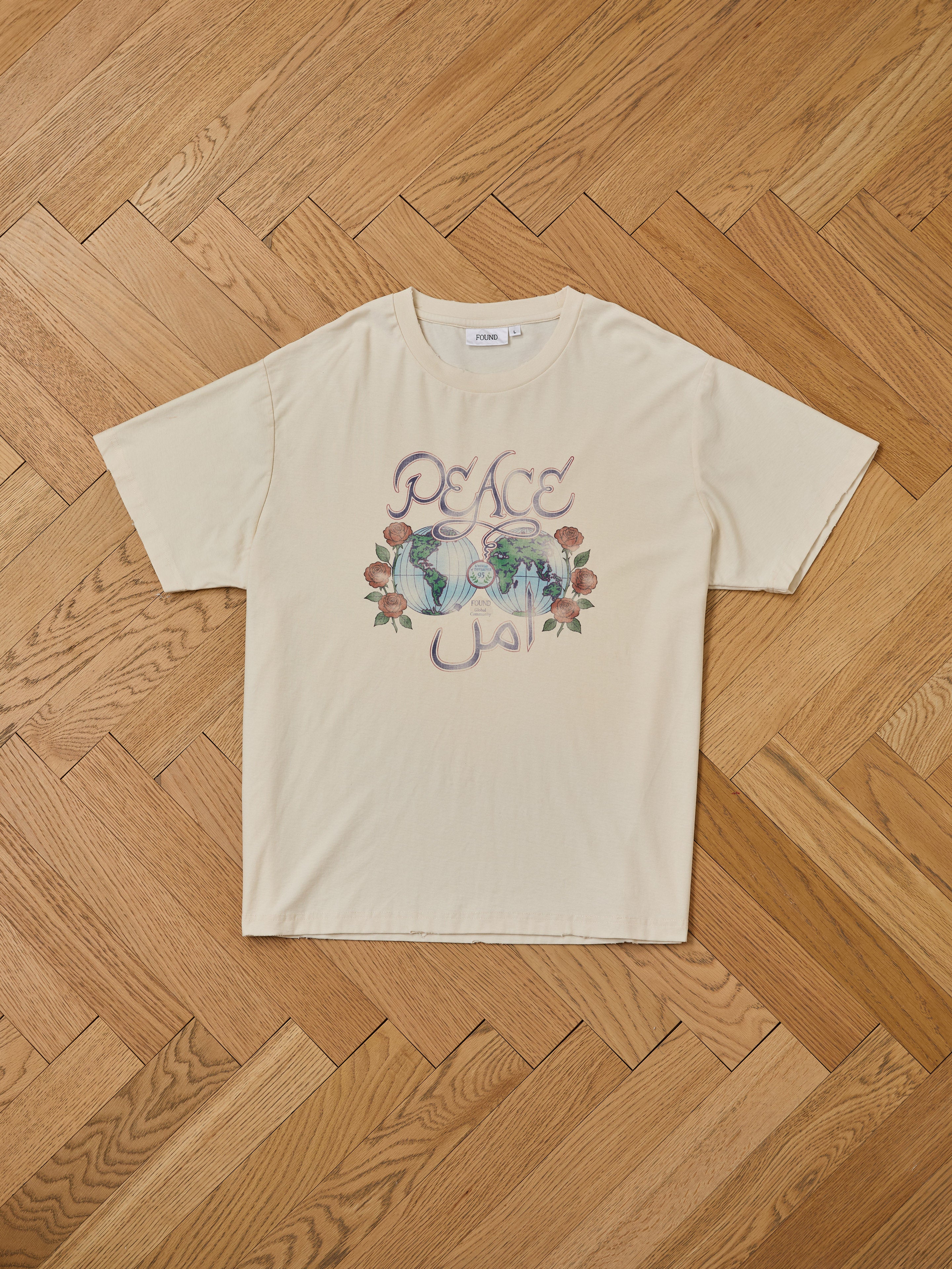 The FOUND Peace Globe Tee, a unisex relaxed fit cotton shirt, lies flat on a wooden floor and displays “PEACE” with its Arabic translation, two illustrated figures, and floral accents.