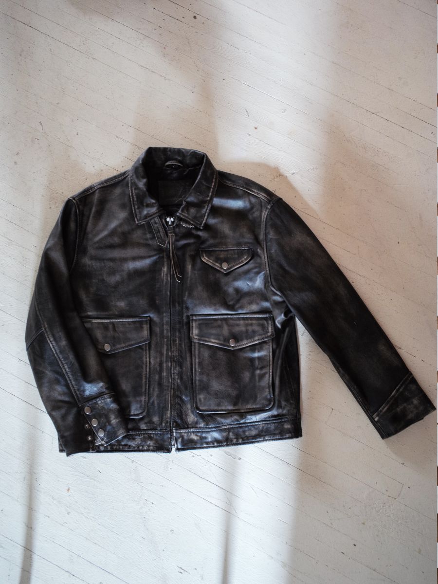 The Distressed Leather Pocket Jacket by Found, a black unisex design crafted from genuine cowhide leather, showcases a front zipper, two large pockets, and a stylish collar. It is elegantly displayed on a light-colored wood surface.