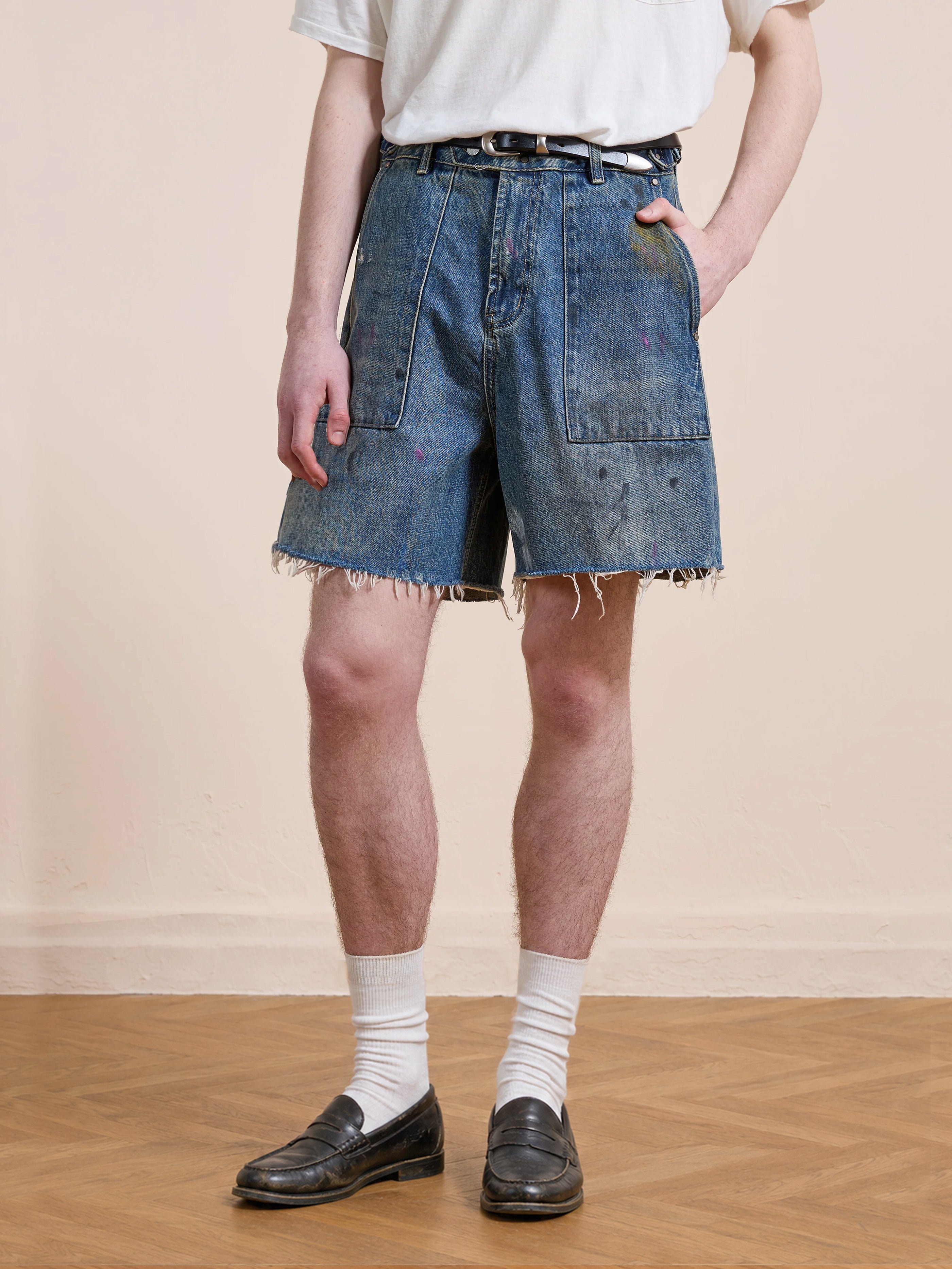 A person wears Found's Paint Studio Denim Shorts—vintage blue wash with paint splatter and frayed hems—paired with a white T-shirt, white socks, and black loafers, standing on a wooden floor against a plain wall.