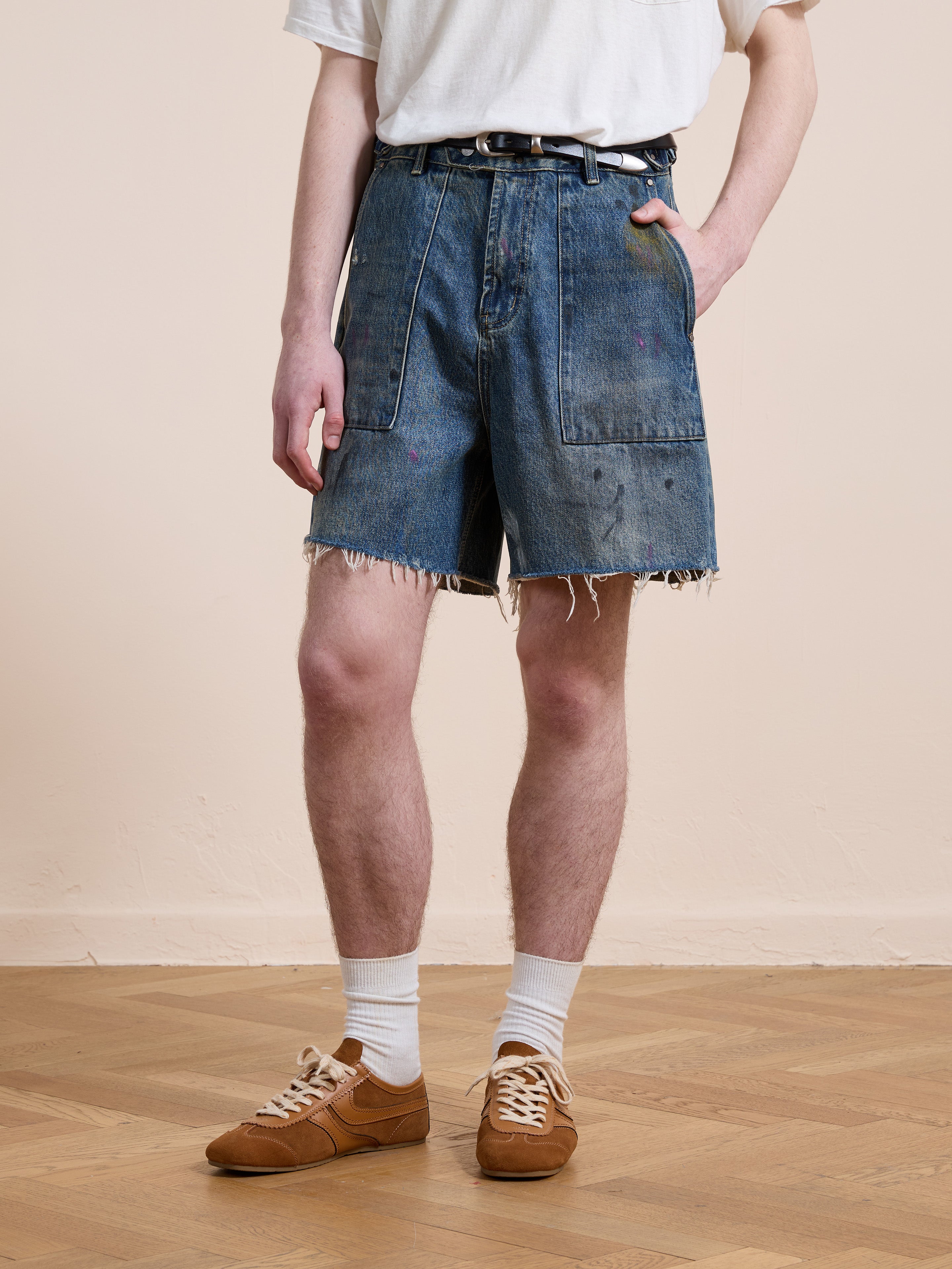 Someone wearing a white t-shirt, Found Paint Studio Denim Shorts, white socks, and brown shoes stands on a wooden floor against a plain wall.