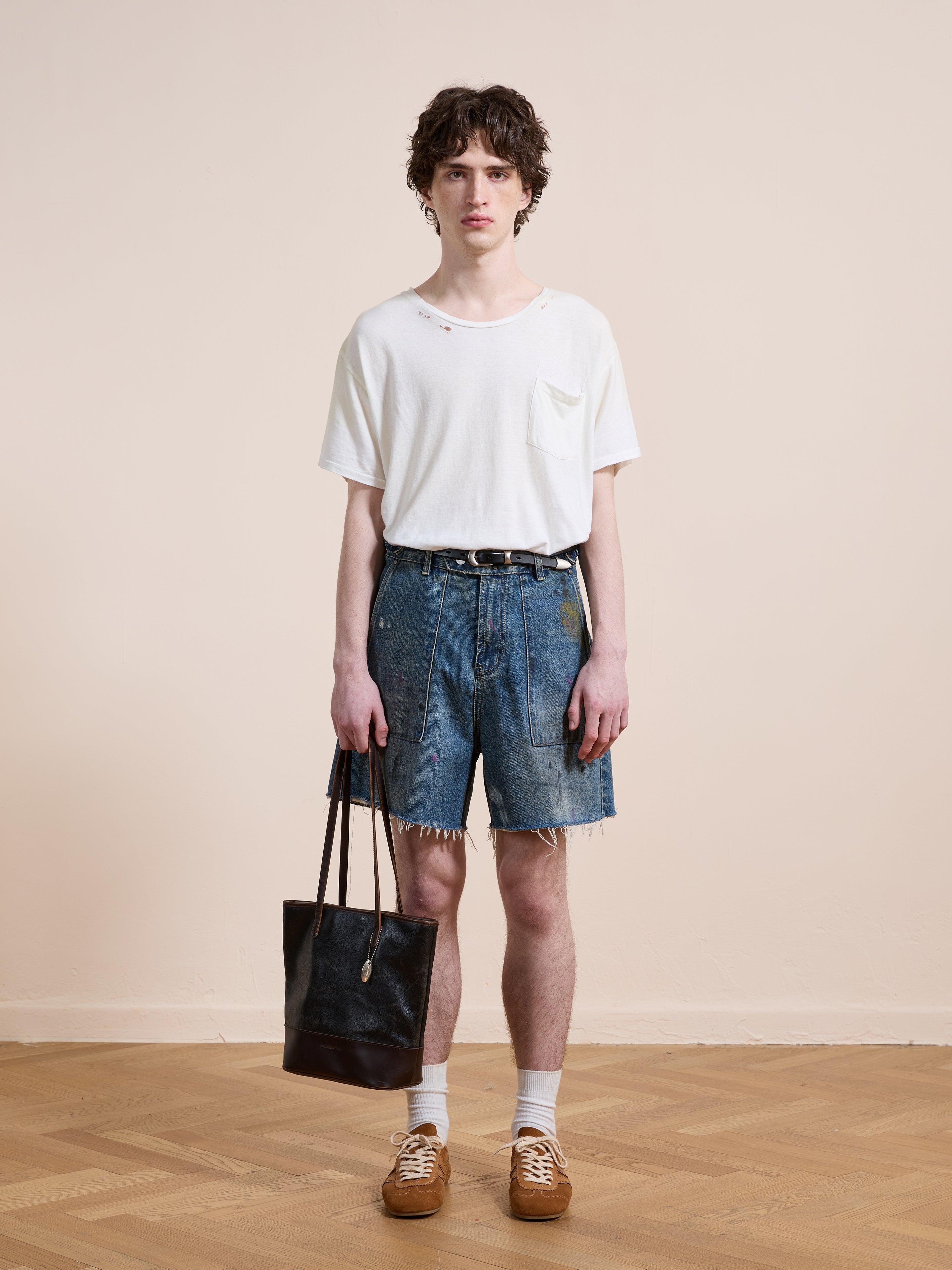A person stands indoors against a plain background wearing a white t-shirt, Found Paint Studio Denim Shorts, white socks, brown shoes, and holding a black tote bag.