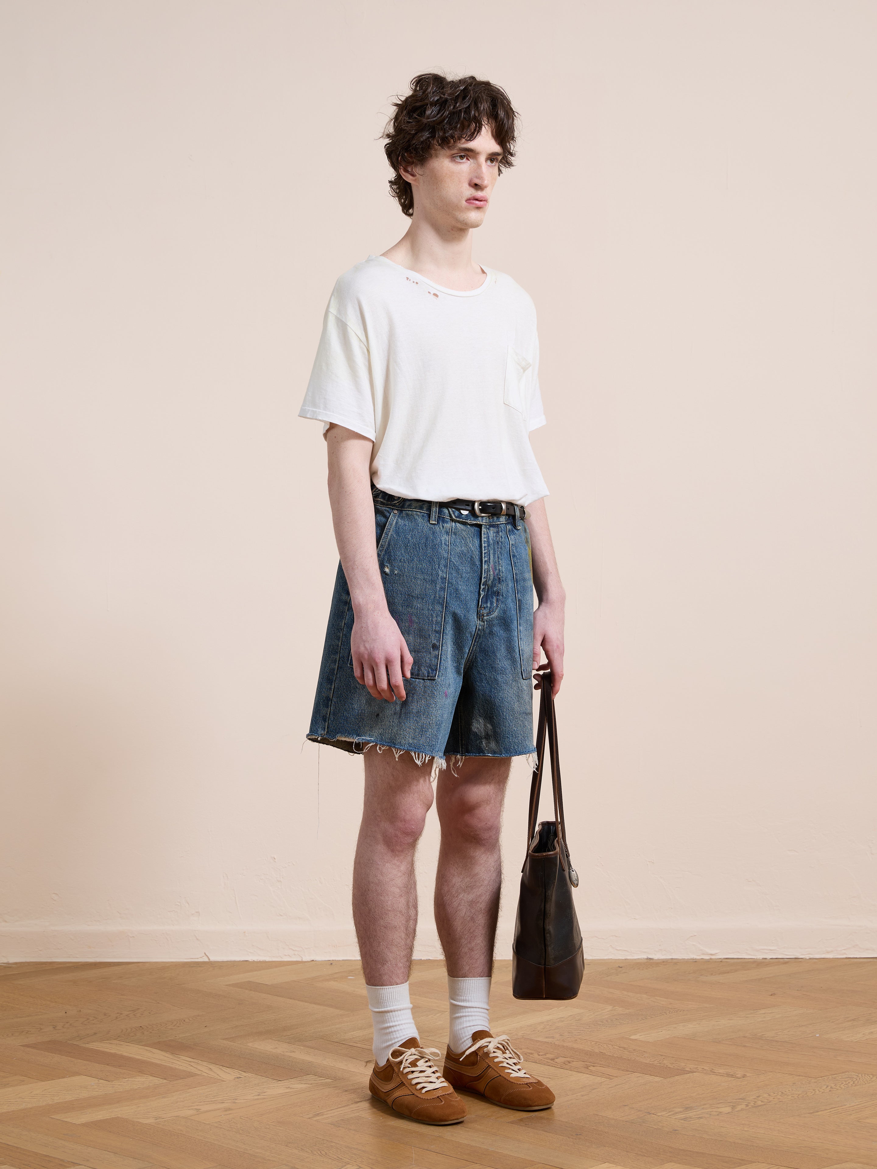 Indoors against a plain beige background, a person wears a white t-shirt, Found Paint Studio Denim Shorts, white socks, brown shoes, and holds a dark handbag.