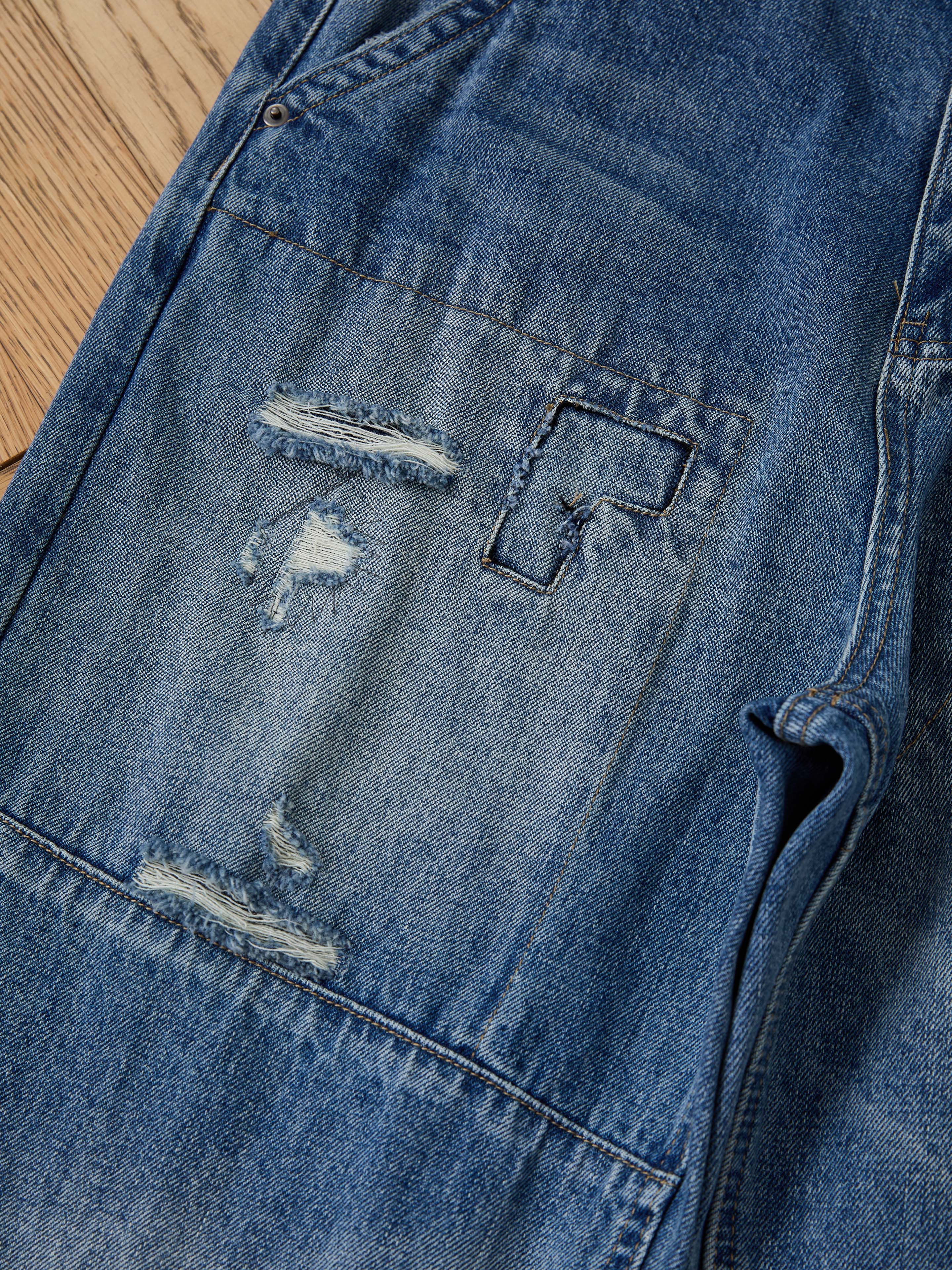 Close-up of FOUND Painter Repaired Jeans, highlighting vintage denim with a ripped, frayed patch and visible mending stitches near the front pocket, displayed on a wooden surface.