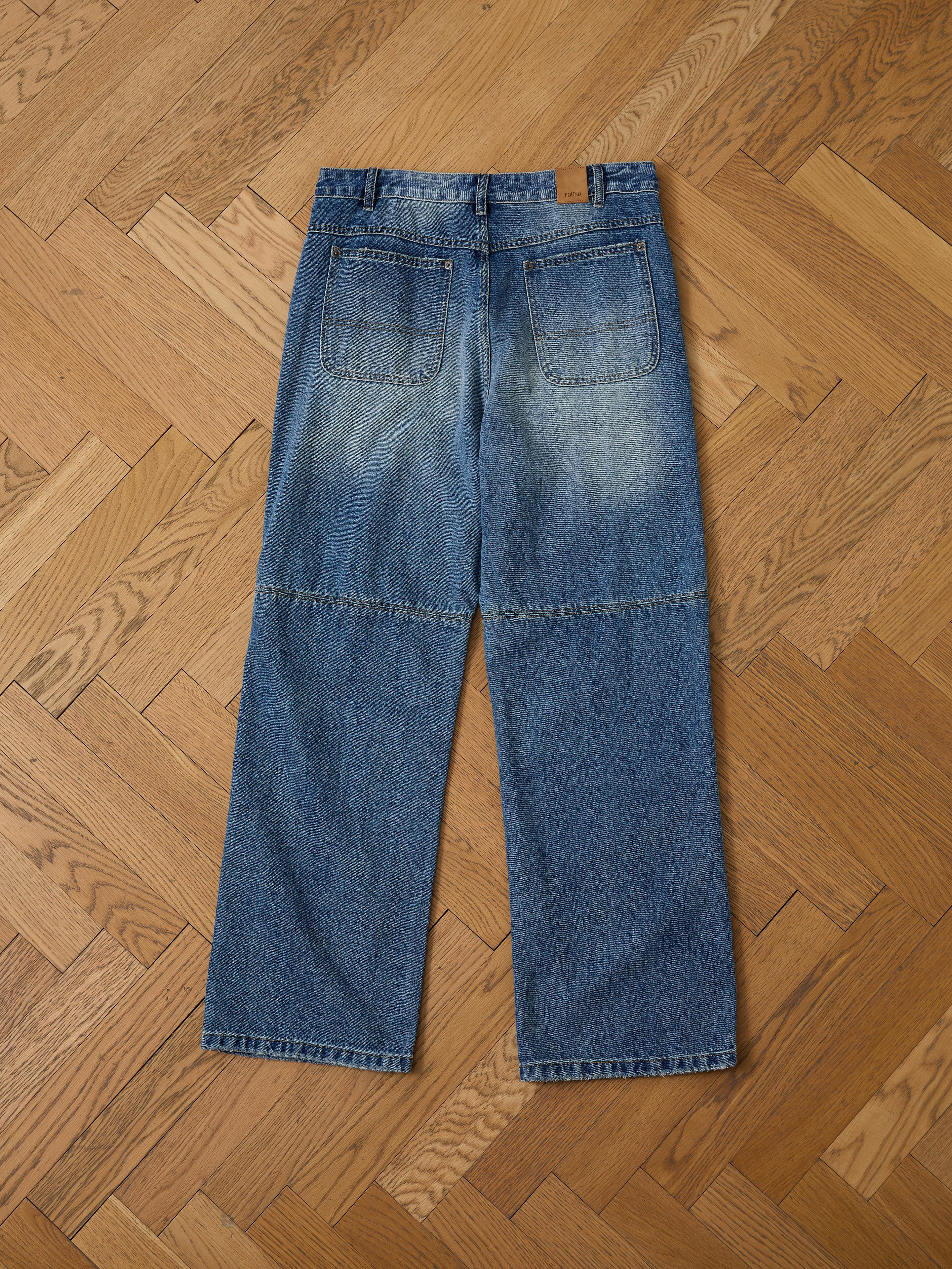 The FOUND Painter Repaired Jeans feature back pockets and visible horizontal fading, displayed flat on a wooden parquet floor.