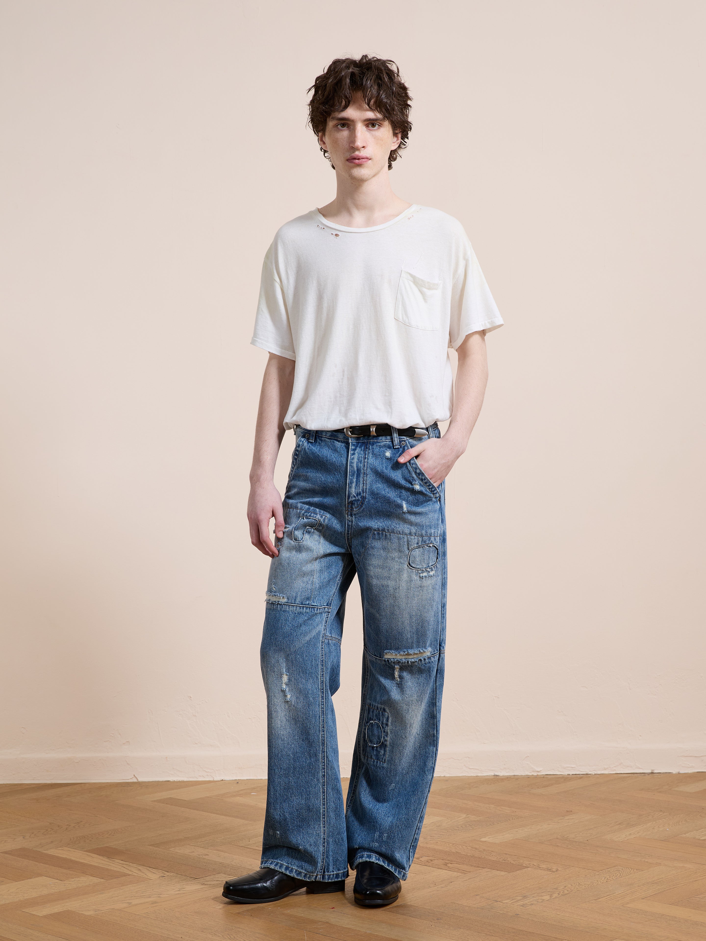 A person stands indoors on a wooden floor against a beige wall, wearing FOUND Painter Repaired Jeans with wide legs, a loose white pocket t-shirt, and black shoes.