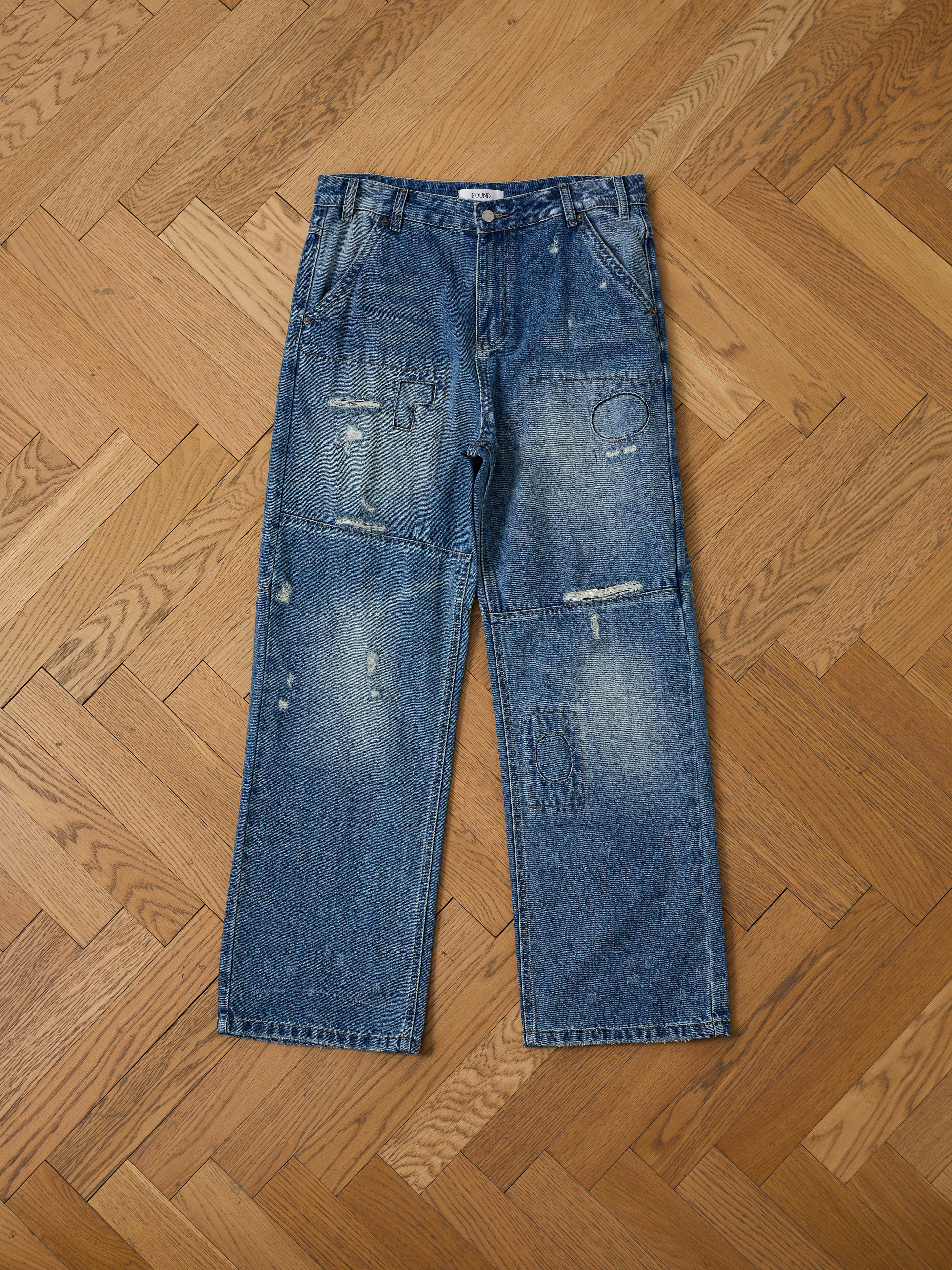 FOUND Painter Repaired Jeans, featuring visible patches and distressed vintage denim, are shown laid flat on a wooden herringbone floor.