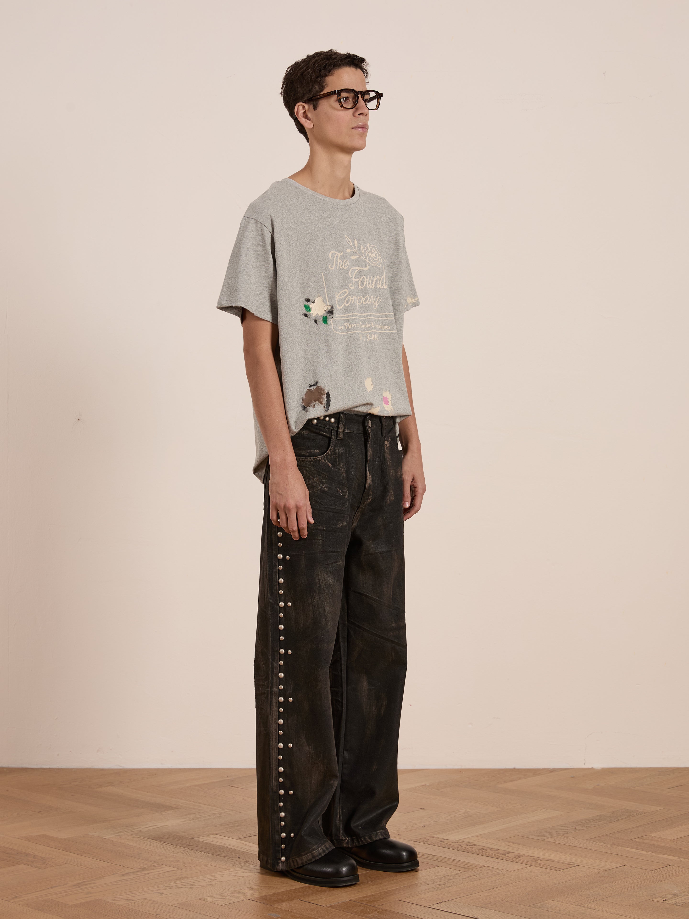 A person in a FOUND Painters Company Tee, featuring a vintage style, pairs it with distressed black studded pants. They're standing on a wooden floor in front of a plain beige backdrop, while the unisex look subtly channels workwear-inspired fashion.
